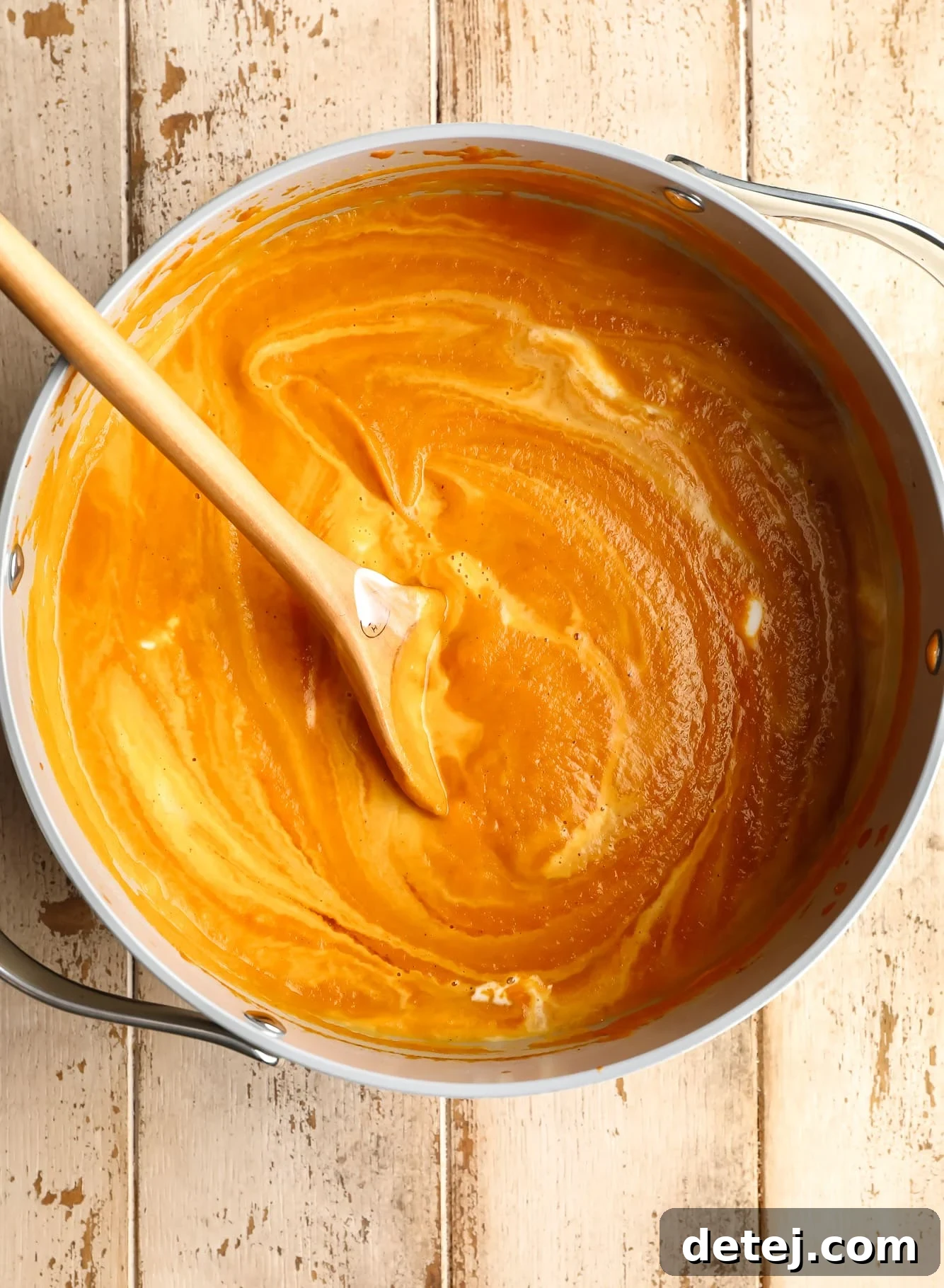 A wooden spoon stirring a large batch of rich, orange vegan pumpkin soup in a grey pot after blending, showing its creamy consistency.