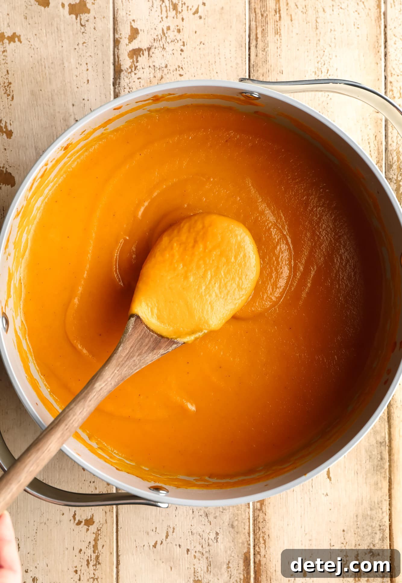 A wooden spoon lifting a spoonful of thick, creamy vegan pumpkin soup from a large grey pot, showcasing its smooth texture.