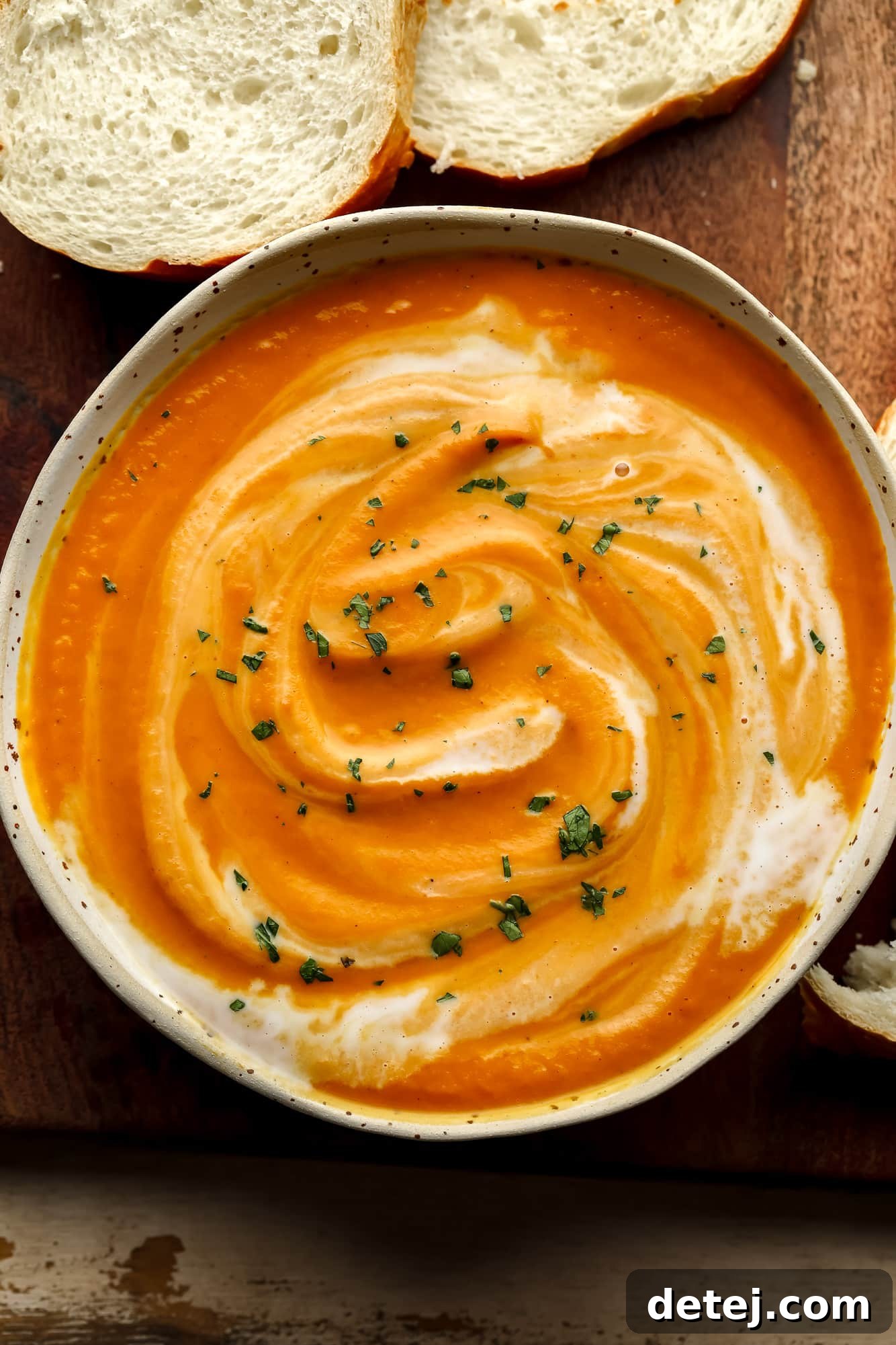 A beautifully presented bowl of creamy vegan pumpkin soup, garnished with a swirl and fresh herbs, surrounded by slices of rustic bread on a wooden board.