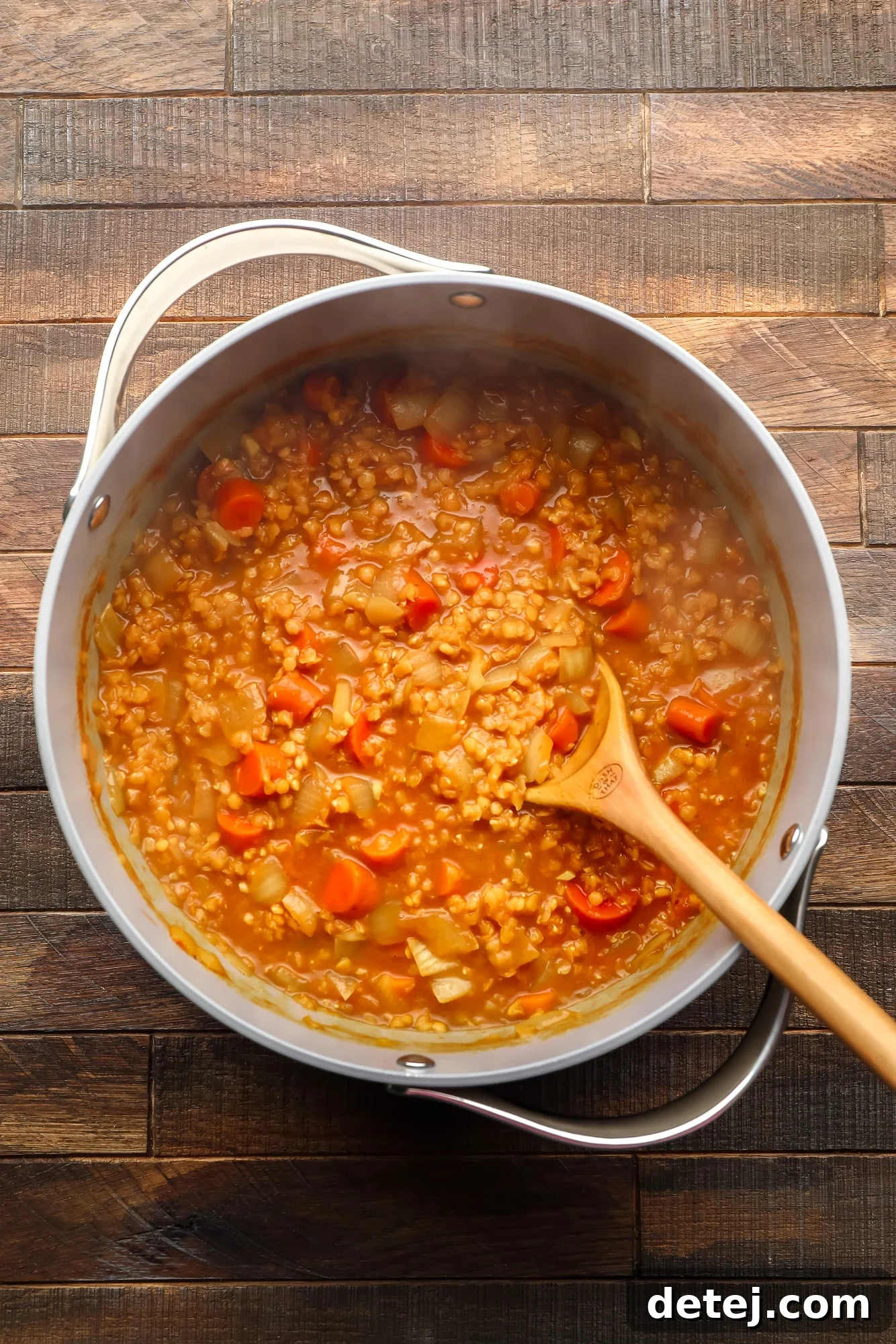 using a wooden spoon to stir carrot and lentil soup in a large grey pot.
