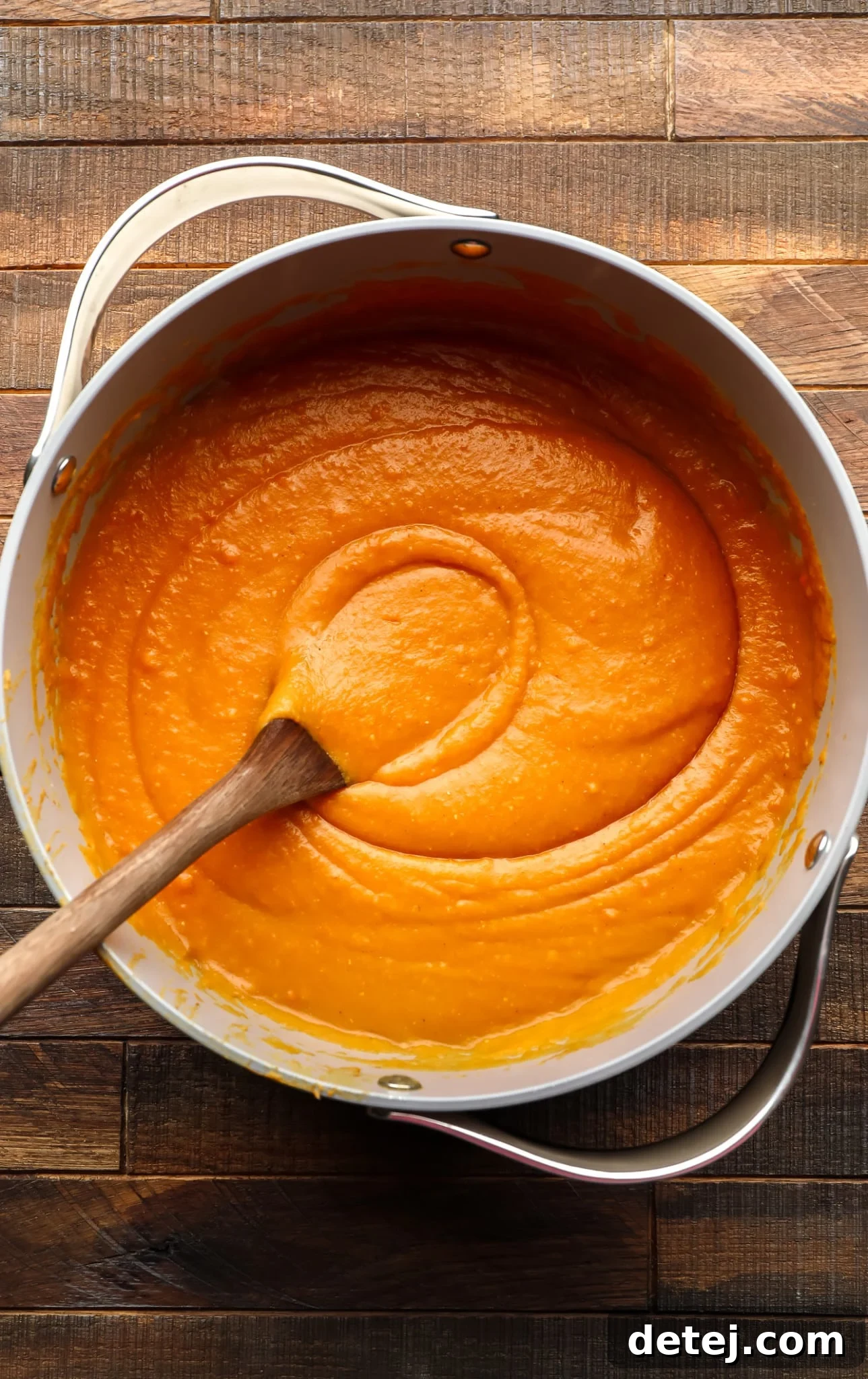 using a wooden spoon to stir creamy carrot and lentil soup in a large grey pot.