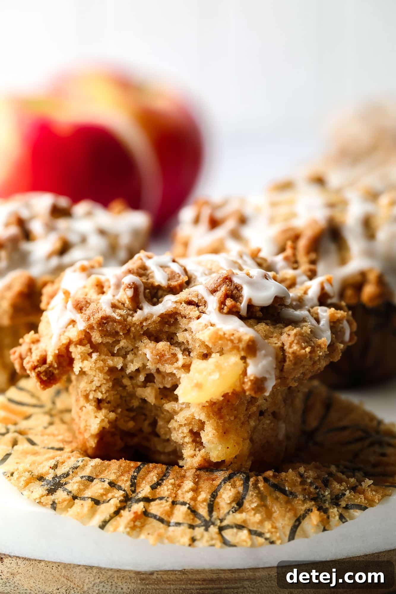 Close up on a vegan apple muffin with a bite taken out, revealing the soft interior, apple chunks, golden crumb topping, and white vanilla icing.