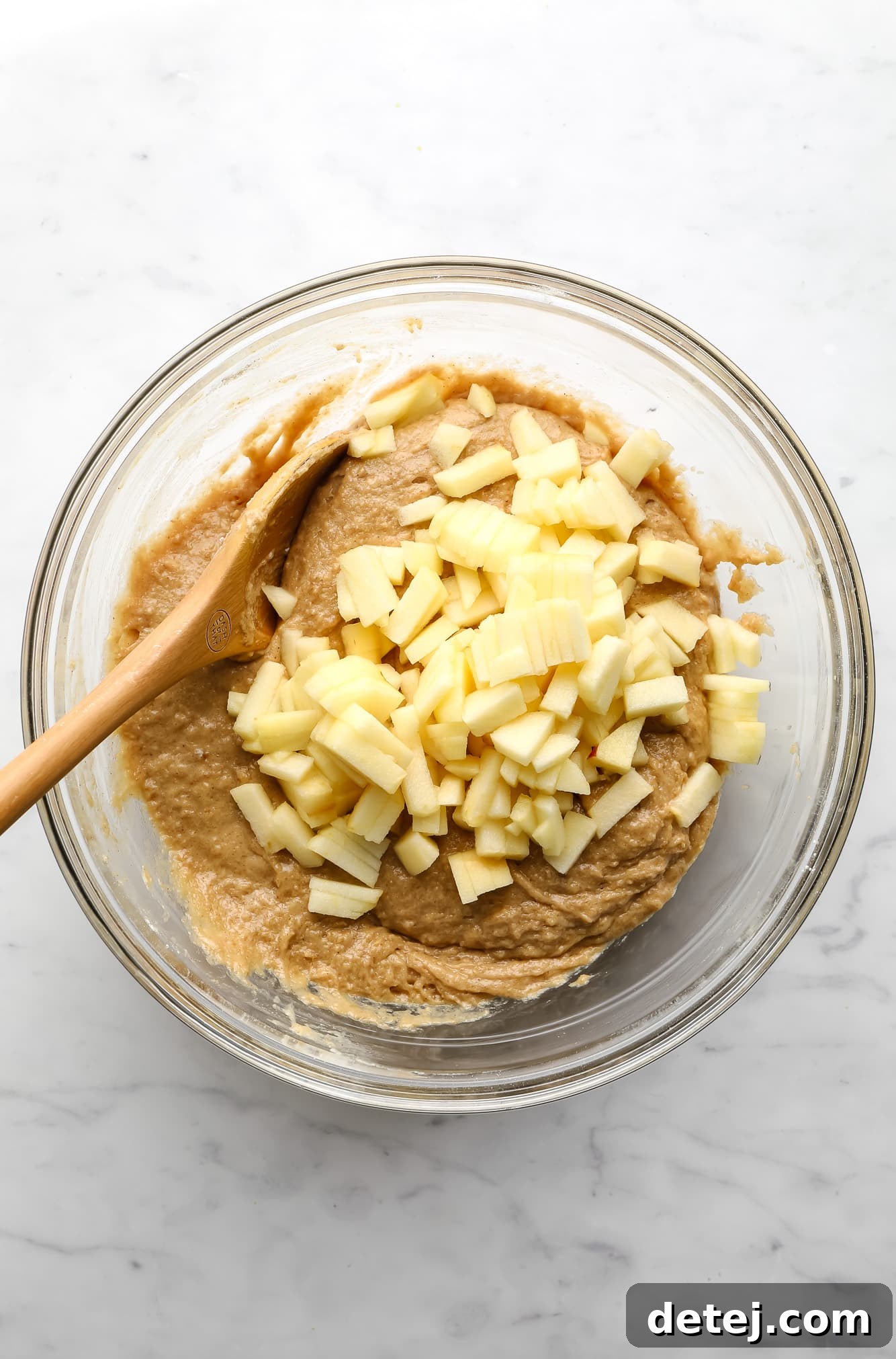 Image showing chopped apples being stirred into vegan apple muffin batter using a wooden spoon in a glass bowl.