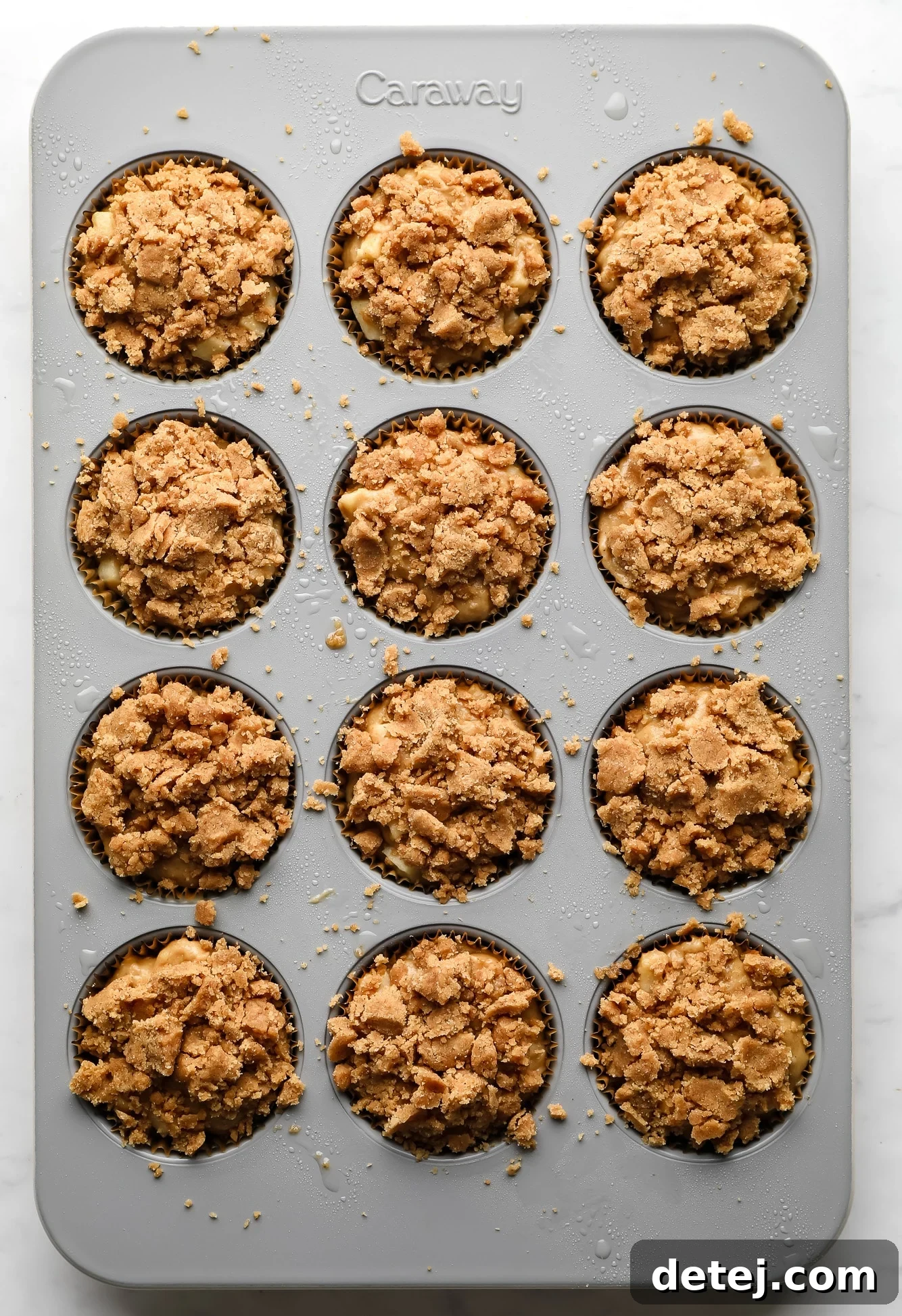 Image of vegan apple muffins topped with a crumble streusel topping in a muffin tin, ready to be baked.