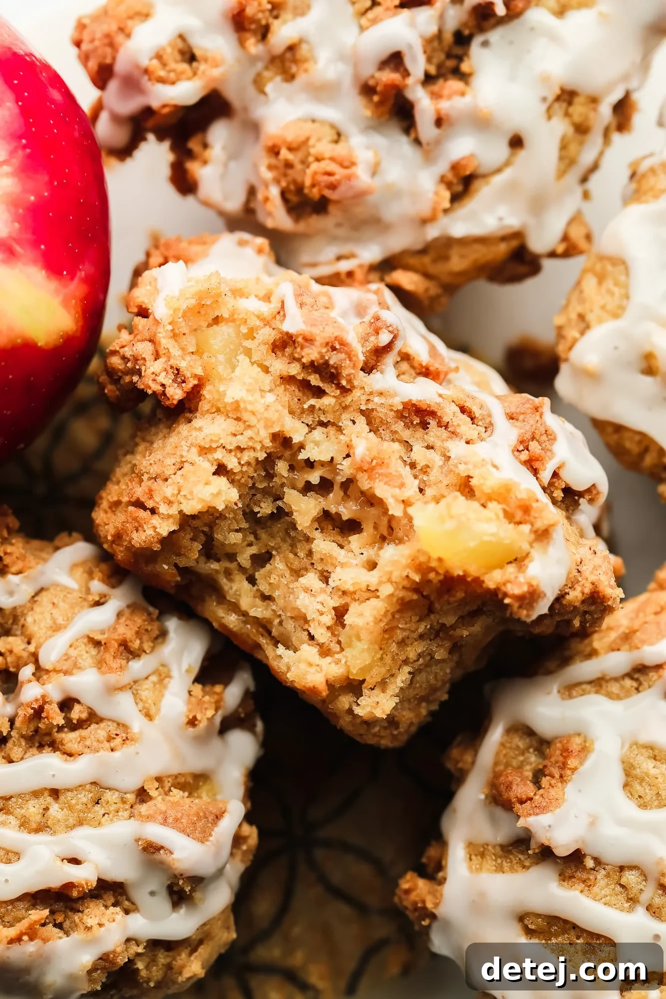 Close up on a vegan apple muffin with a bite taken out of it, showcasing the soft, apple-studded interior and the golden, crumbly topping with a vanilla glaze drizzle.