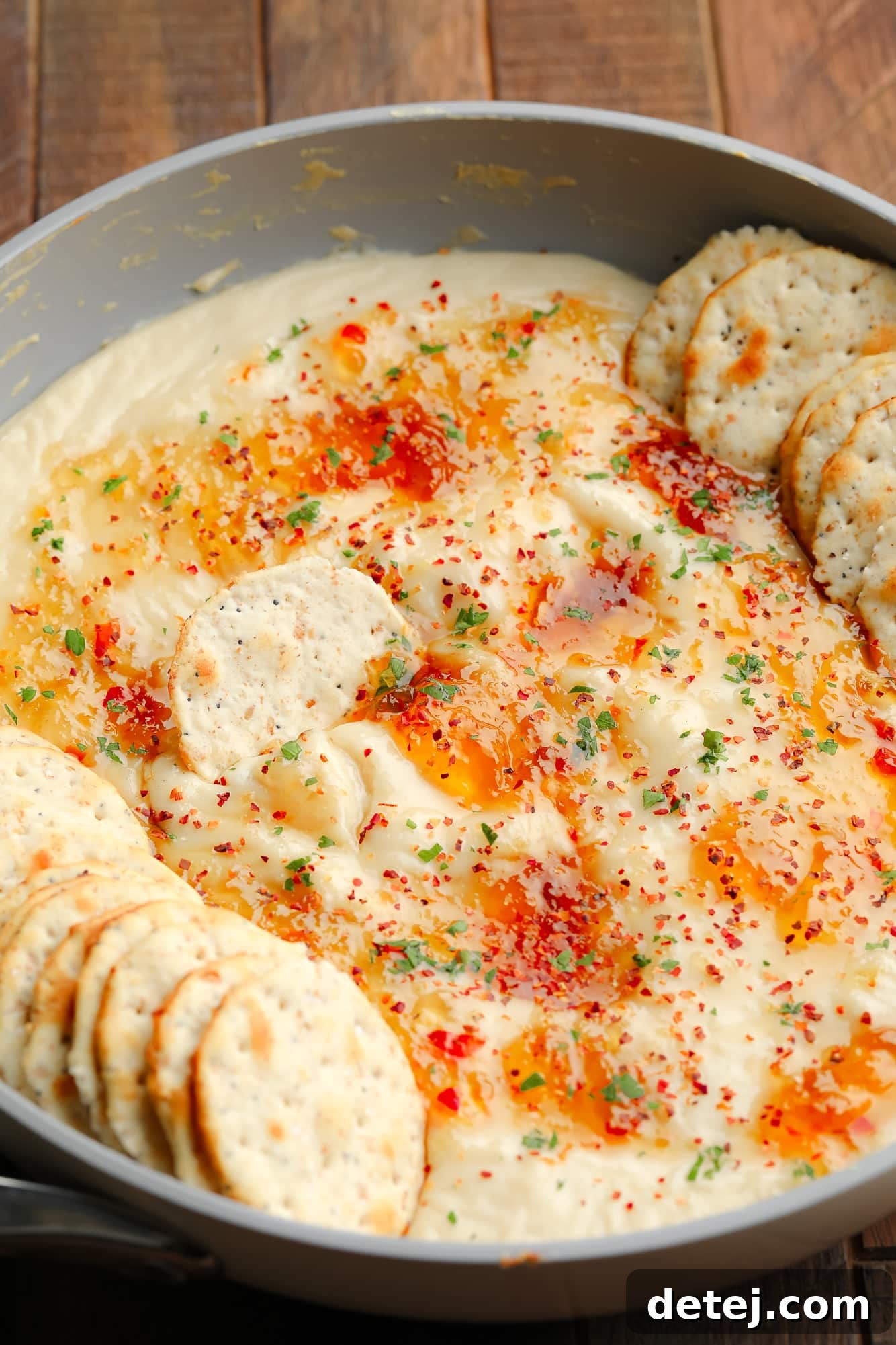 A rustic grey saucepan filled with bubbling, golden-brown baked vegan brie dip, generously topped with glistening red pepper jelly, surrounded by an assortment of crunchy crackers.