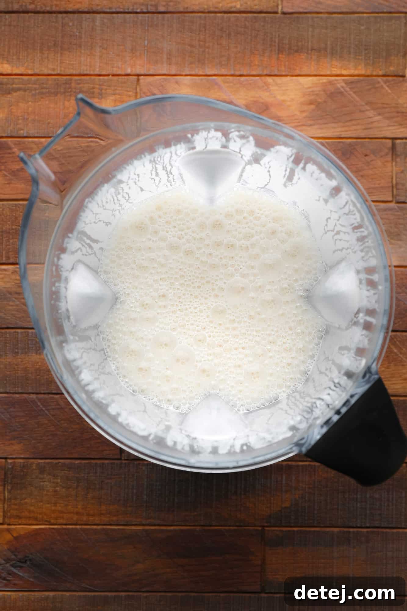 A high-powered blender contains a smooth, off-white, slightly bubbly liquid mixture, ready to be transformed into creamy vegan brie.