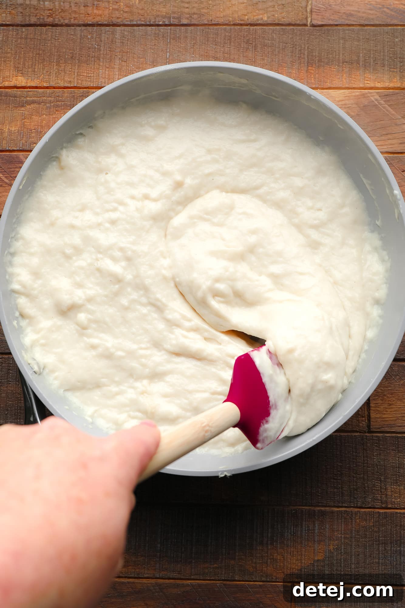A red silicone spatula is actively stirring a creamy, white, thick vegan brie cheese mixture in a grey oven-safe saucepan on the stovetop.