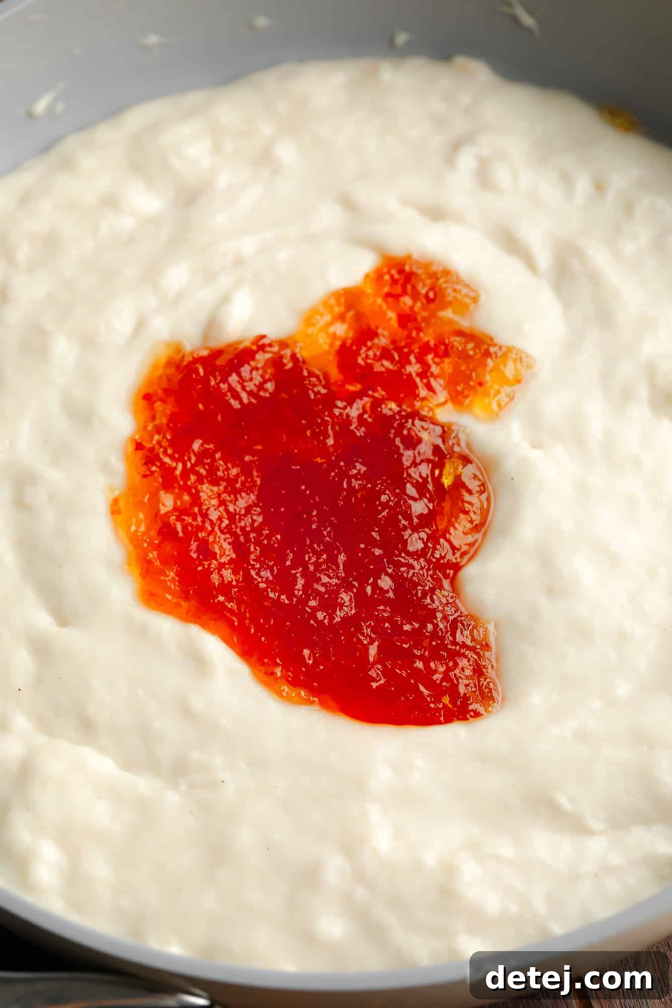 A generous dollop of vibrant red pepper jelly rests atop a creamy, white base of vegan brie cheese in an oven-safe dish, ready for baking.