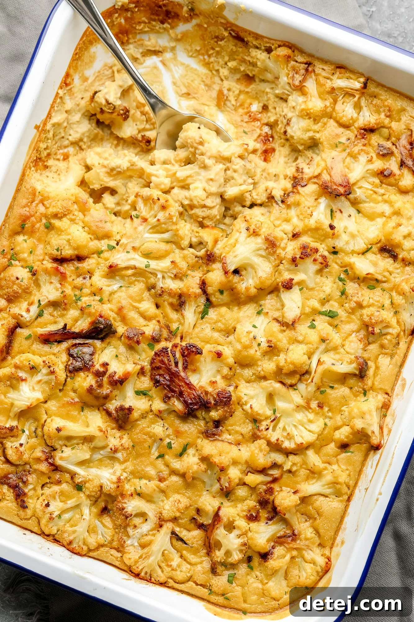 A large metal spoon scoops a generous portion of golden-brown vegan cauliflower cheese from a white baking dish, showcasing its creamy texture and roasted florets.