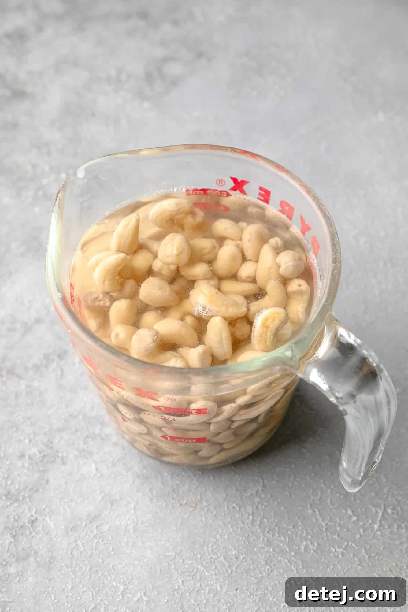 A clear glass measuring cup holds raw cashews submerged in hot water, illustrating the soaking process.