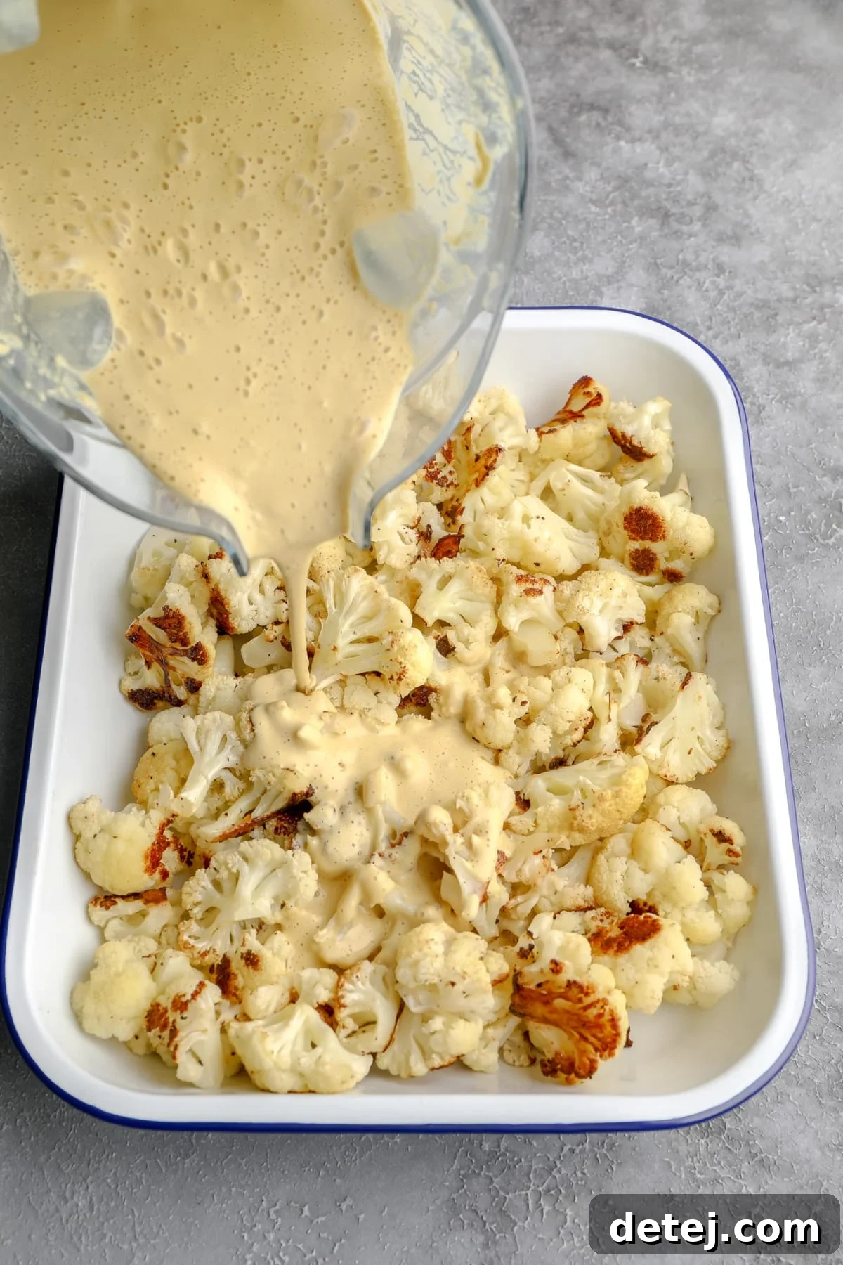 A rich, golden vegan cheese sauce is poured over perfectly roasted cauliflower florets in a large white baking dish, ready for baking.