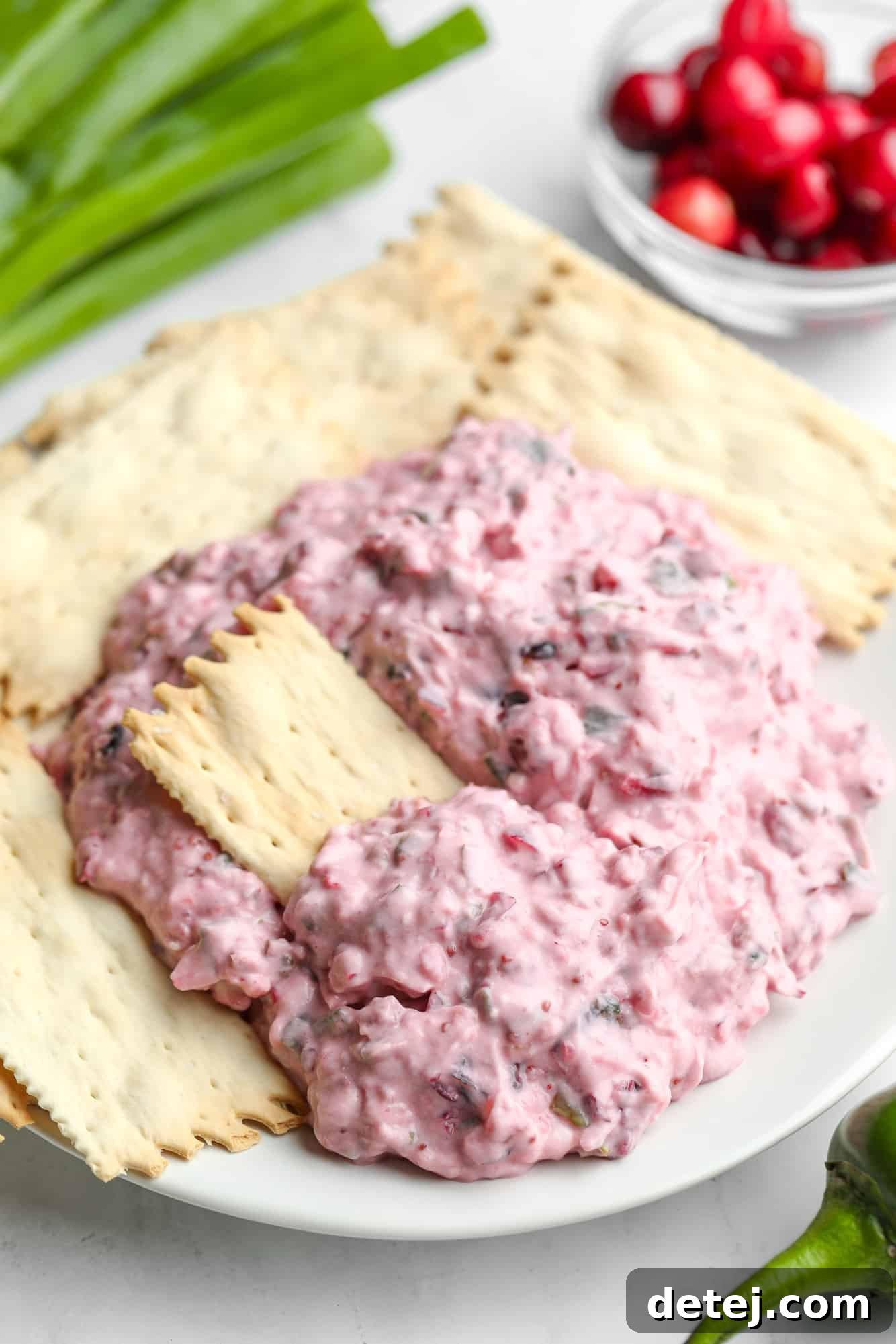A beautifully presented bowl of creamy, pink Cranberry Jalapeno Dip, adorned with fresh cranberries and thinly sliced green onions, surrounded by an assortment of crispy crackers on a pristine white plate, making for an inviting holiday appetizer spread.