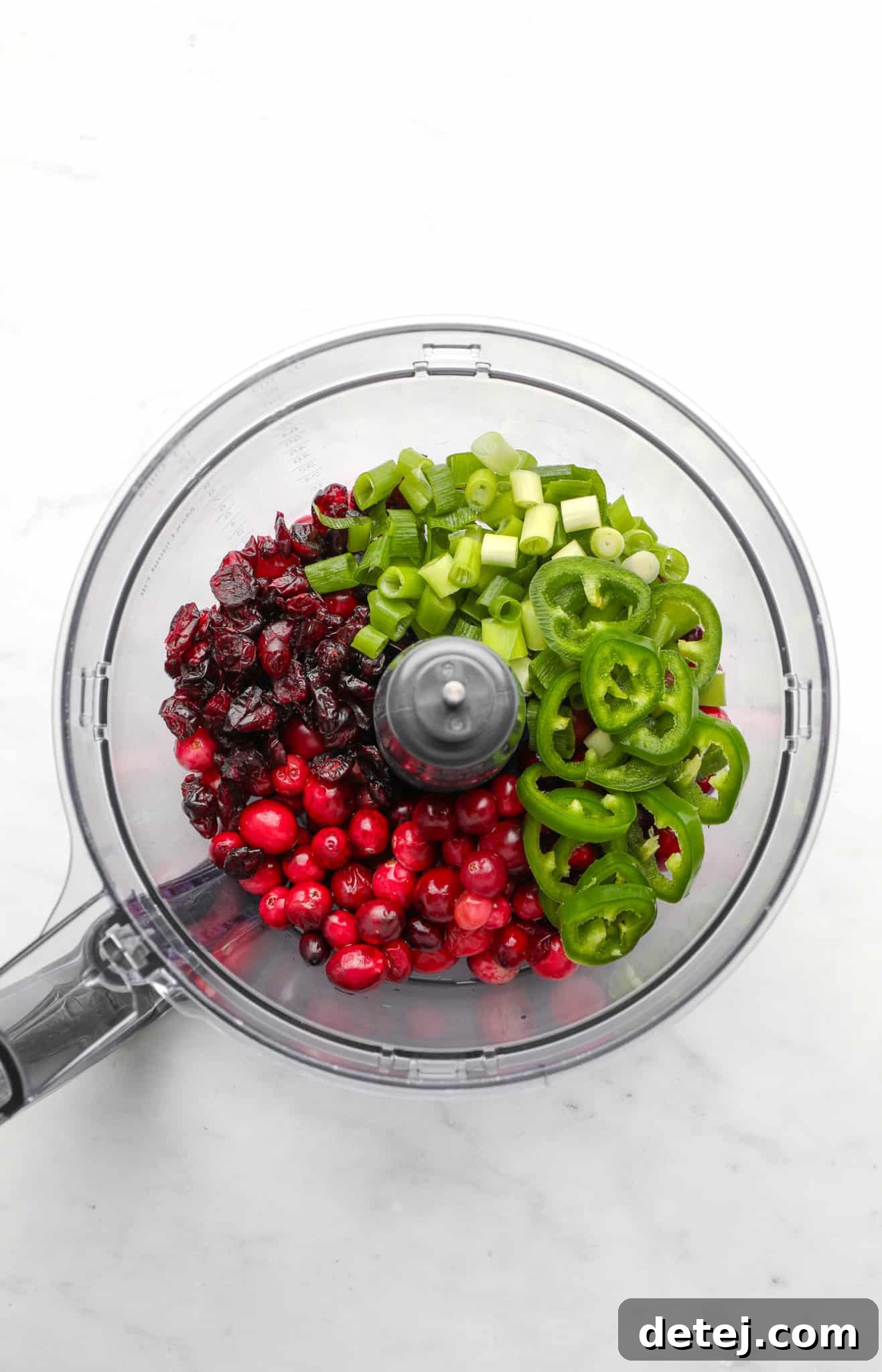 A top-down view of a food processor bowl filled with the colorful raw ingredients for Cranberry Jalapeno Dip: fresh cranberries, dried cranberries, vibrant green onions, and thinly sliced jalapeno pepper, all awaiting processing.