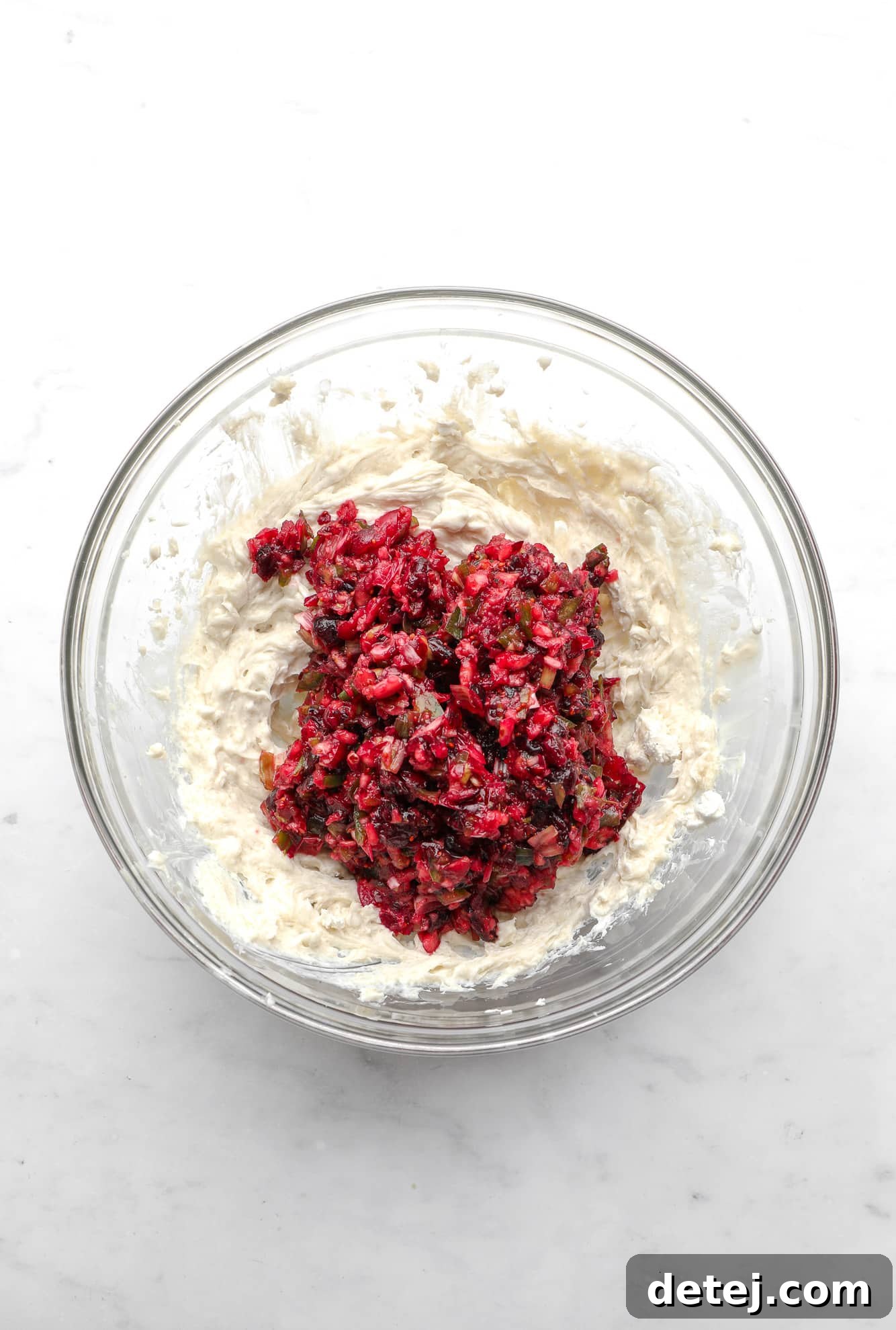 A glass mixing bowl showcases the distinct layers of the Cranberry Jalapeno Dip in progress: a fluffy, whipped vegan cream cheese base at the bottom, topped with the vibrant, finely chopped mixture of cranberries, green onions, and jalapeno, poised for mixing.