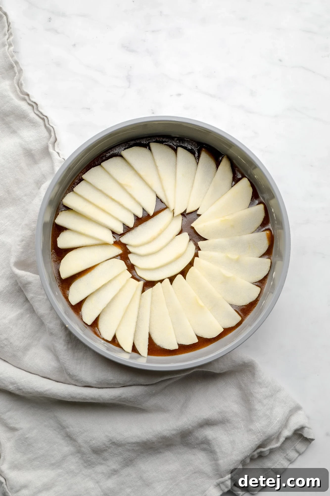 Sliced apples carefully layered on top of the rich vegan caramel sauce in a round grey cake pan.