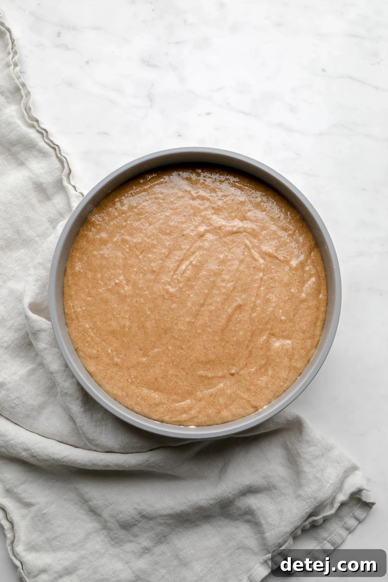 Brown cake batter covering the apple and caramel layers in a round grey cake pan, ready for baking.