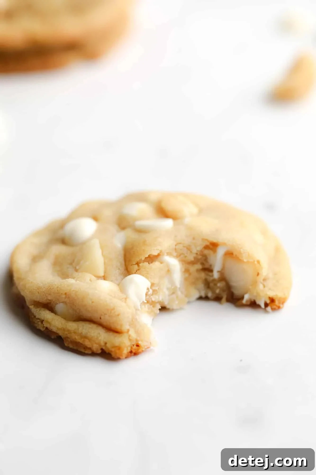Close-up of a delightful Vegan White Chocolate Macadamia Nut Cookie with a bite taken out of it, revealing its rich texture and generous filling of white chocolate and macadamia nuts.