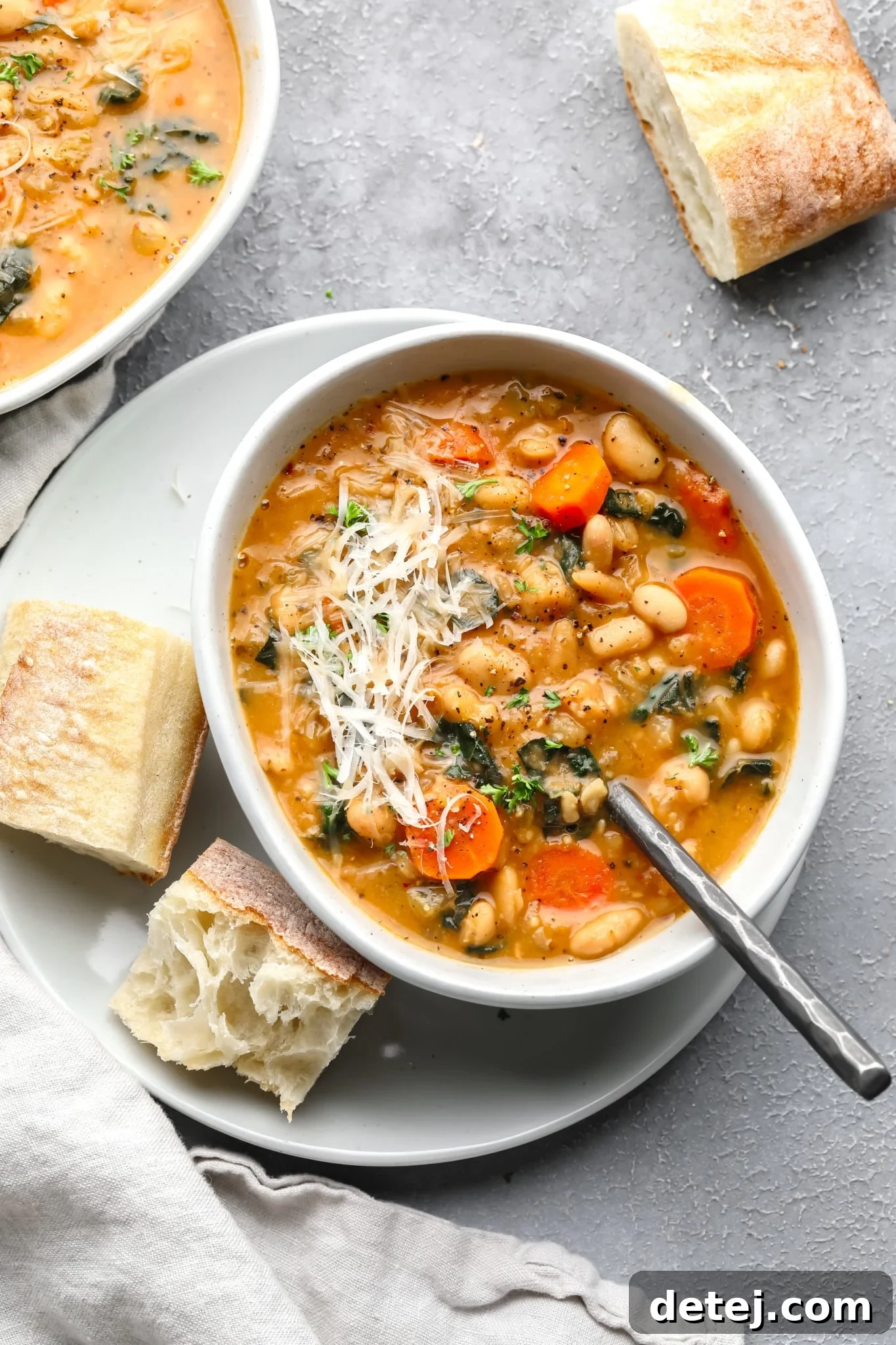 A white bowl of creamy white bean soup topped with vegan parmesan, served with pieces of crusty bread on the side.