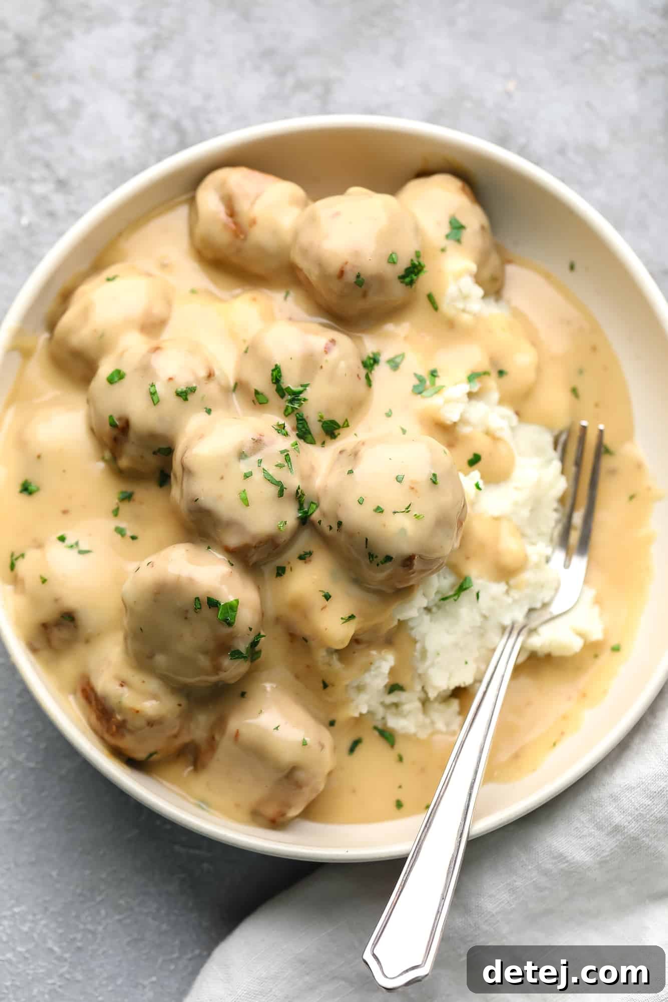 Vegan Swedish Meatballs generously covered in a luscious, creamy gravy and served on a bed of fluffy mashed potatoes in a pristine white bowl, accompanied by a fork.