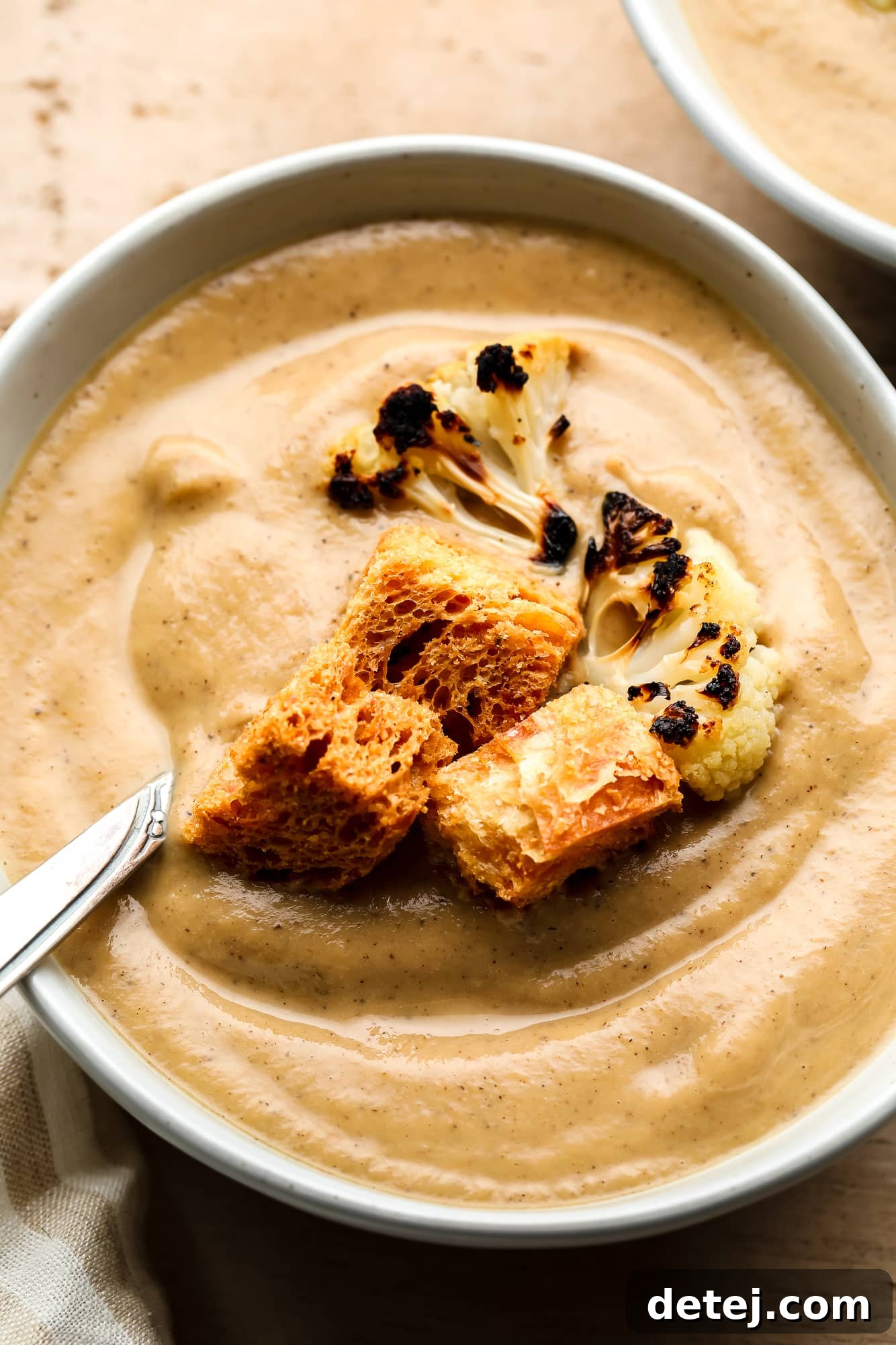 Close-up of luxuriously creamy roasted cauliflower soup, garnished with golden croutons and perfectly charred cauliflower florets, served in an elegant white bowl.