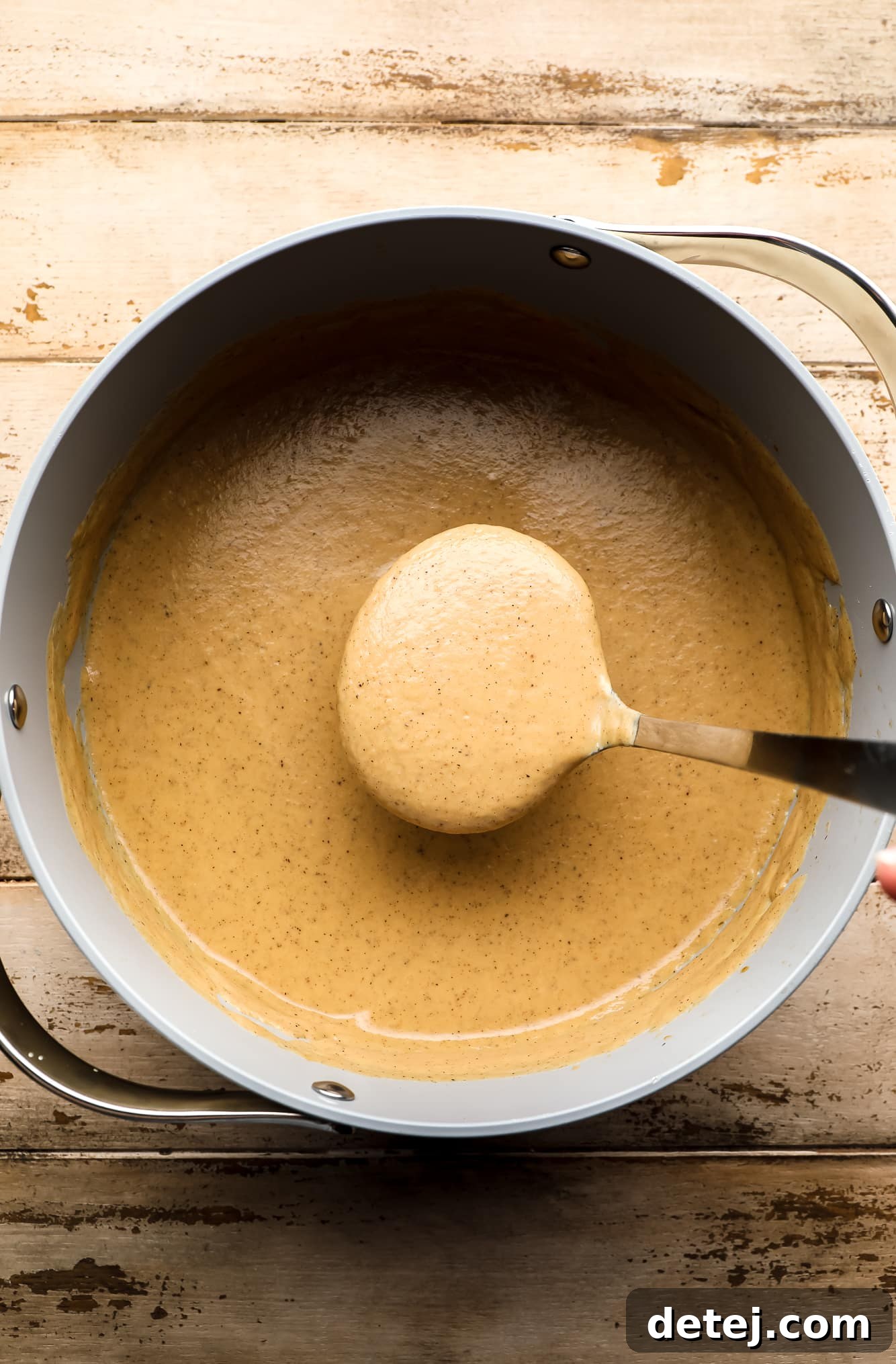 A ladle gracefully lifting a generous scoop of the creamy roasted cauliflower soup from a large batch simmering in a grey pot, showcasing its rich texture.