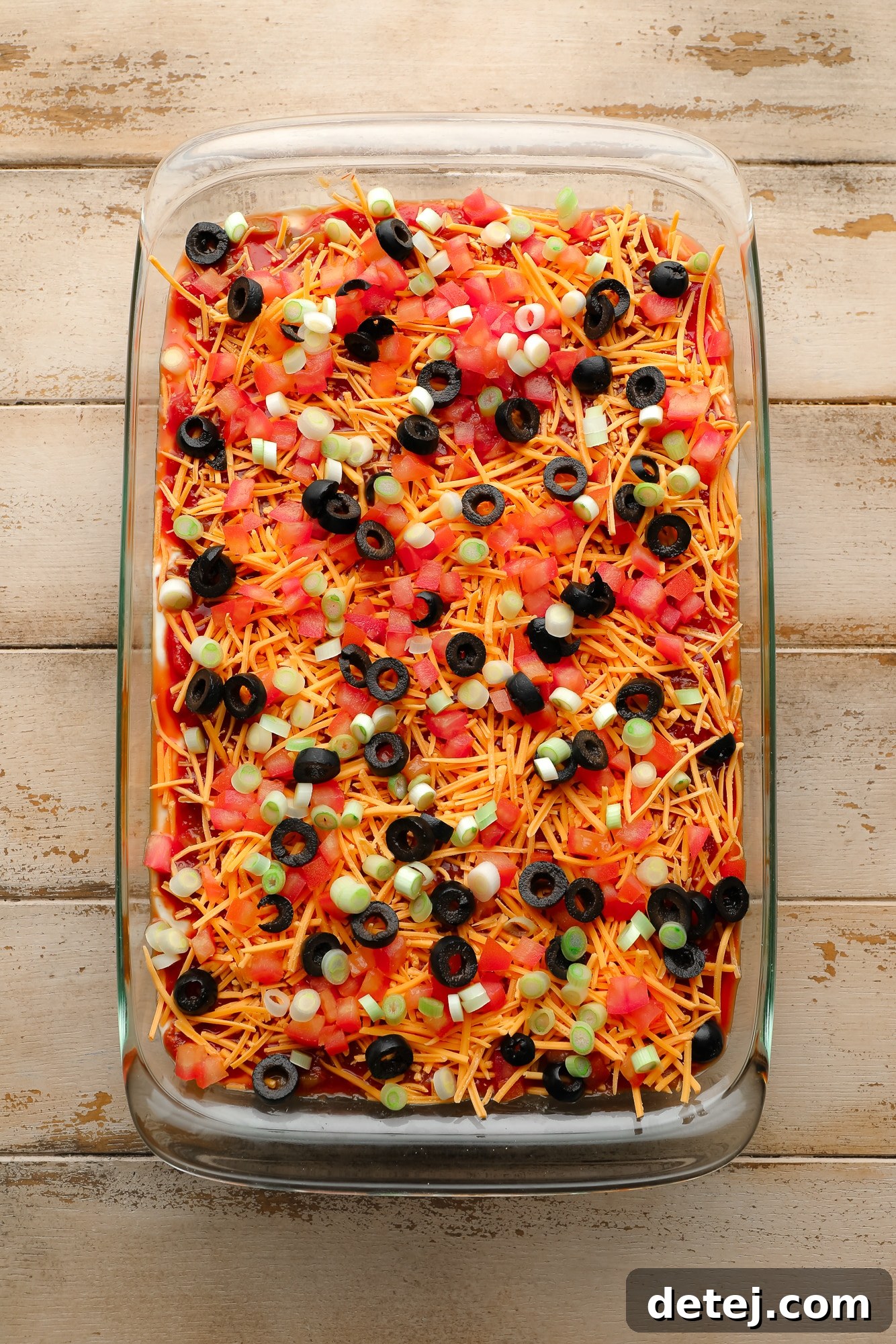 Vegan 7 Layer Dip topped with black olives, shredded vegan cheese, and diced tomatoes in a glass casserole dish.