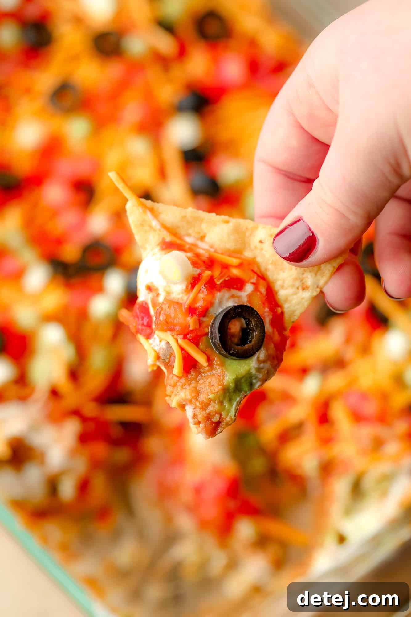 womans hand holding a tortilla chip that's been dunked in vegan 7 layer dip.