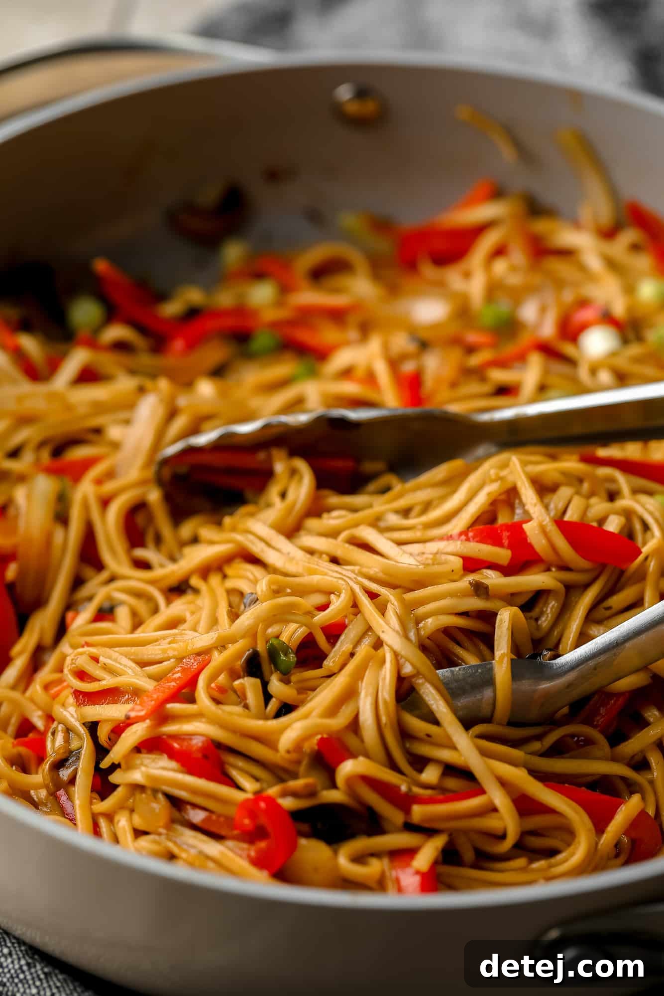Metal tongs serving a portion of vegan lo mein from a large gray skillet, highlighting the delicious blend of noodles and vegetables.