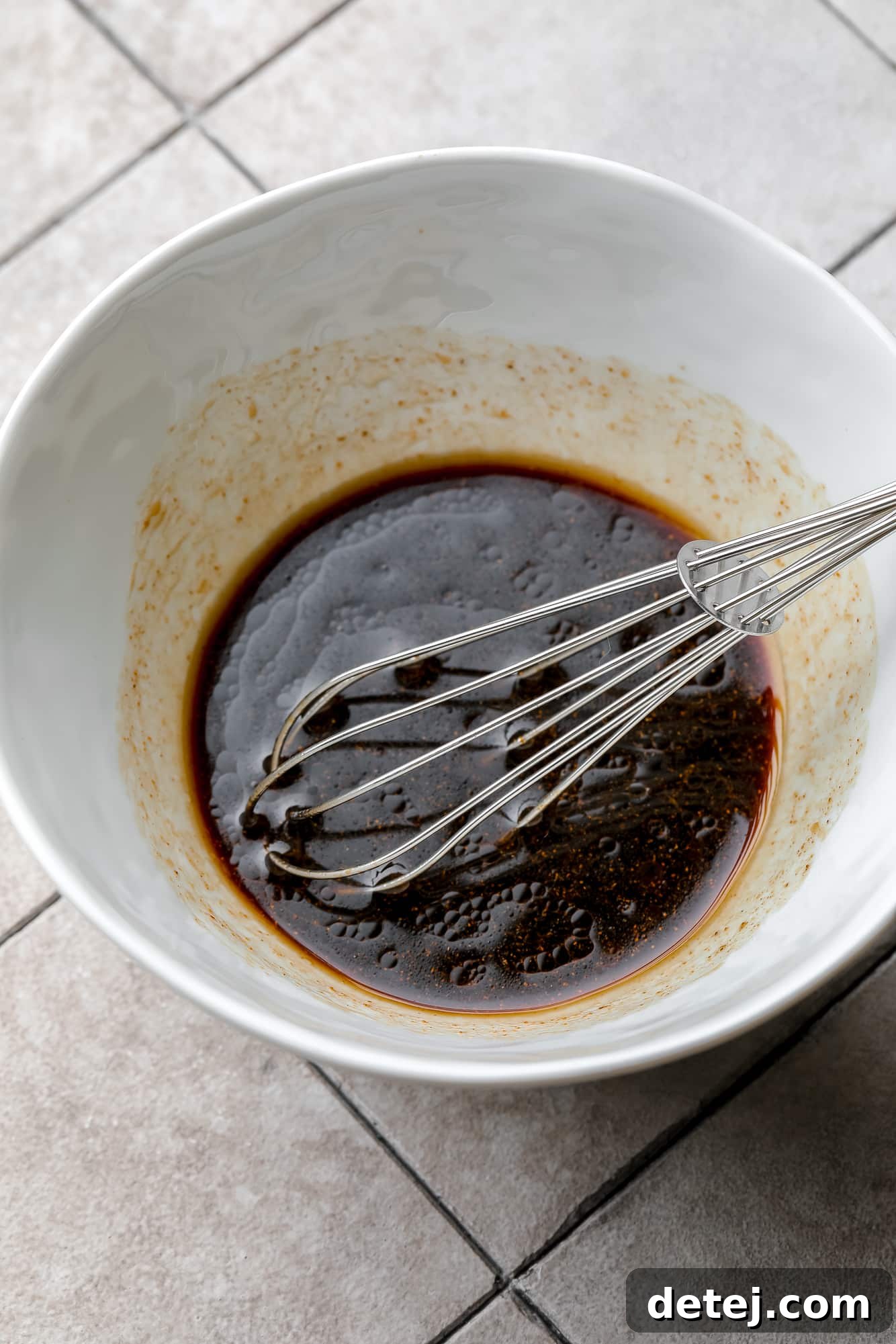 A close-up view of a hand whisking homemade vegan lo mein sauce in a white bowl, showcasing its smooth consistency.