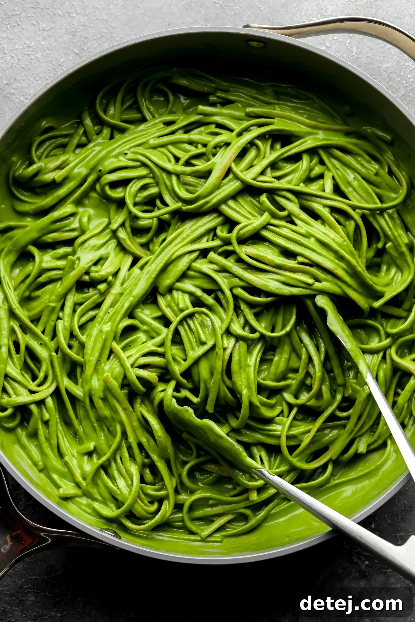 using metal tongs to toss long pasta noodles in a bright green sauce in a large grey pot.