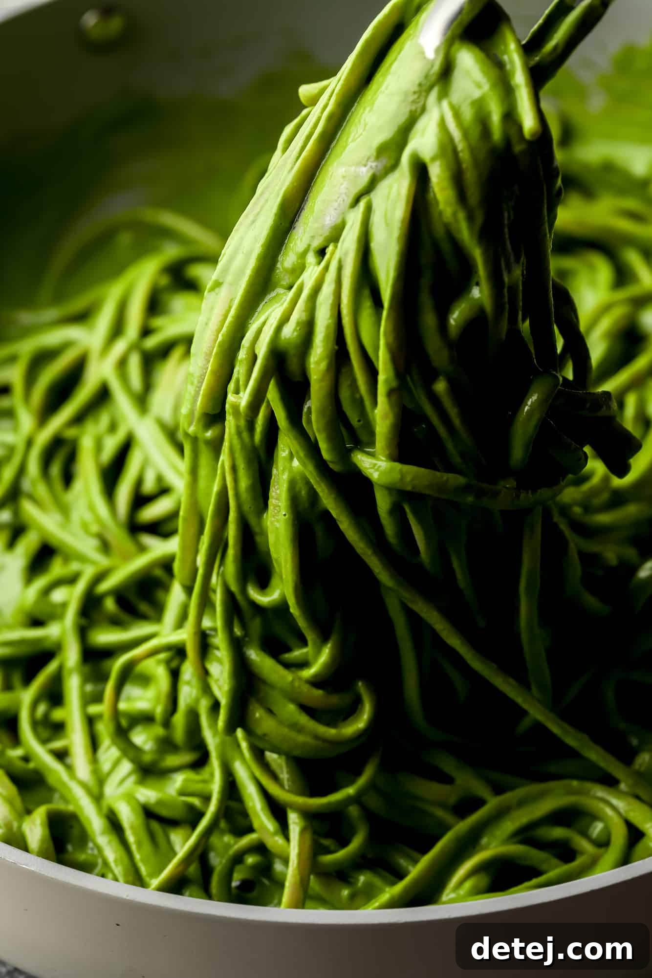 metal tongs lifting a scoop of green pasta from a large pot.