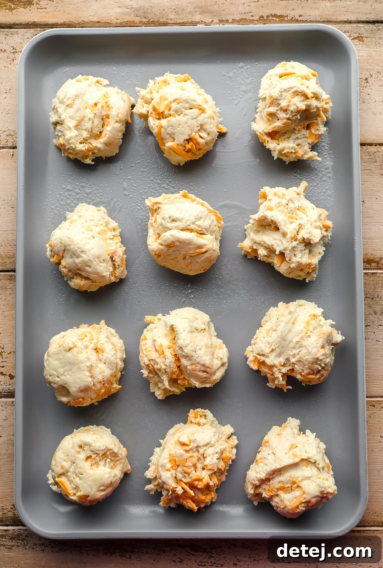 Scoops of raw vegan cheddar bay biscuit dough, perfectly portioned and arranged on a metal baking sheet, ready for the oven.
