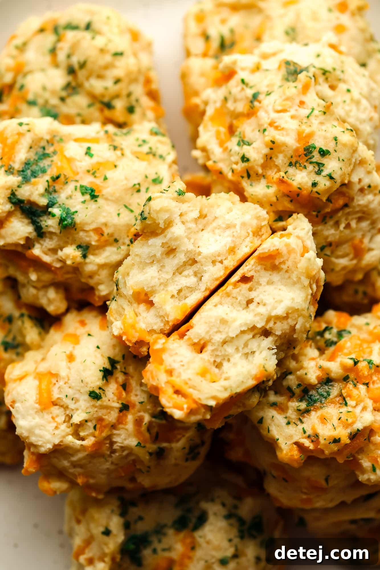 A vegan cheddar bay biscuit torn in half, revealing its fluffy interior, with a pile of whole biscuits in the background.