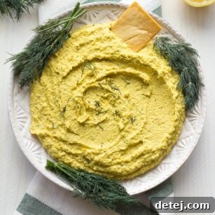 Bright yellow lemon dill hummus in a bowl, garnished with fresh dill sprigs, served with pita chips and cucumber slices.