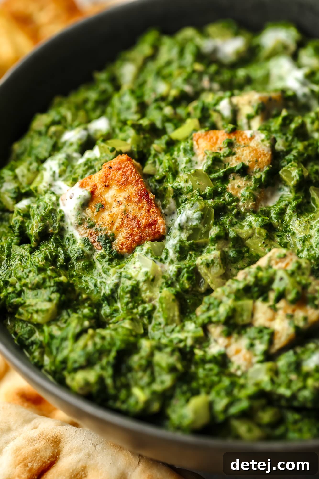 Close up on golden tofu cubes tossed in a batch of vegan palak paneer in a black bowl, garnished with fresh cilantro.