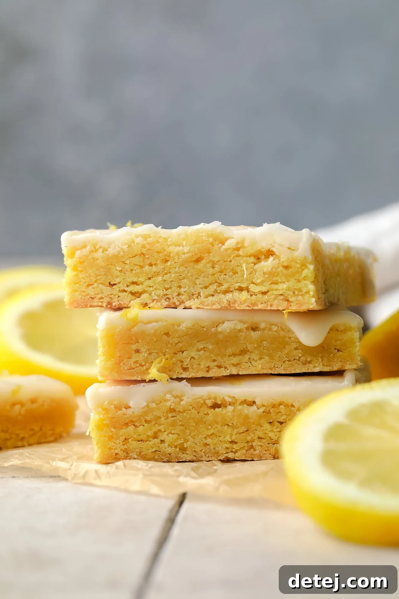 A side view of a stack of golden vegan lemon brownies, each glistening with a smooth lemon glaze, highlighting their perfect thickness and inviting texture.