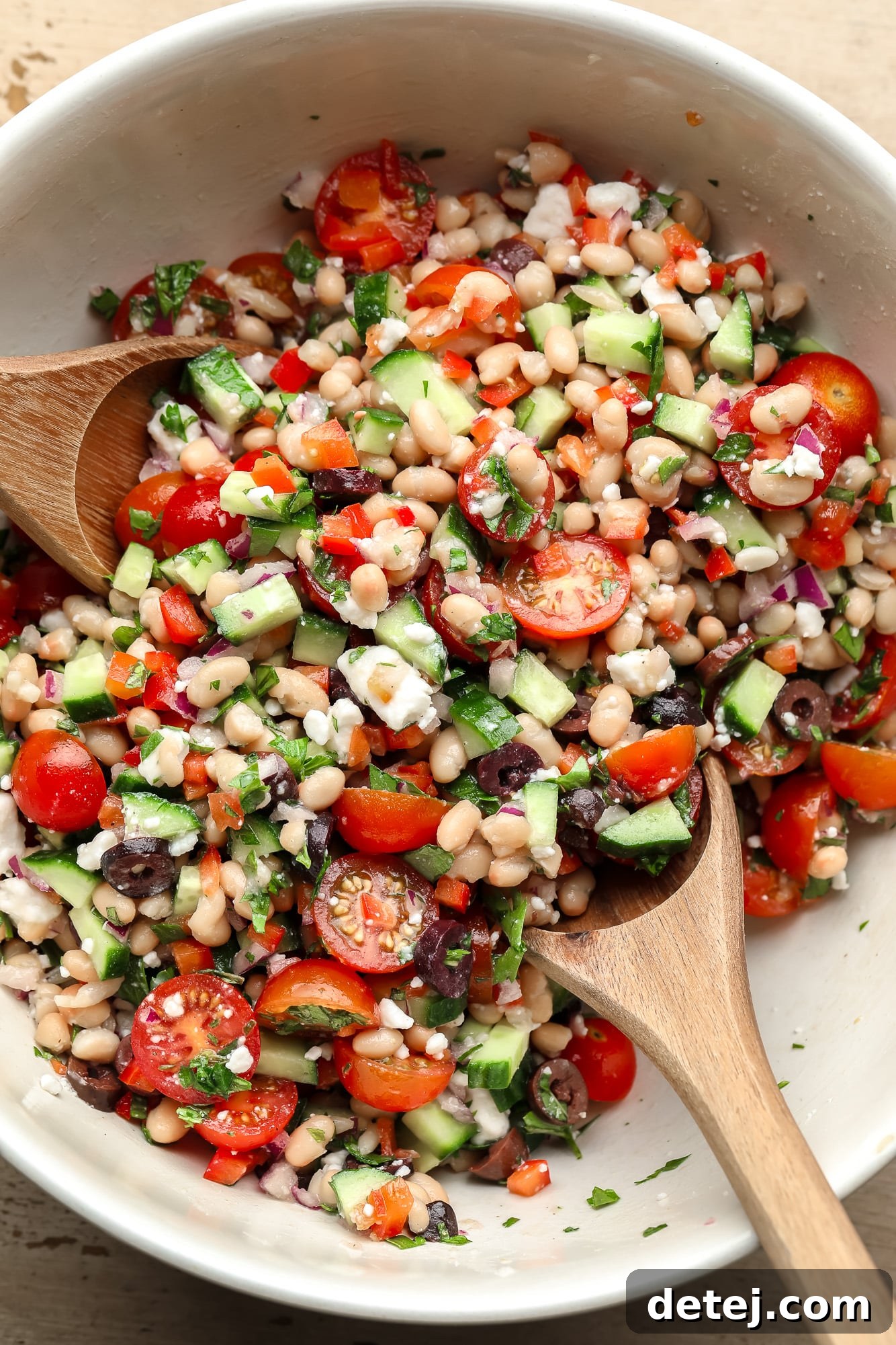 Two wooden spoons gently tossing a vibrant Mediterranean white bean salad in a large white bowl.