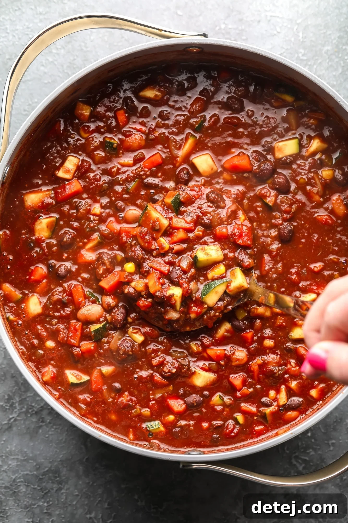 A woman's hand using a wooden spoon to stir a large pot of simmering veggie chili, filled with colorful vegetables and beans.