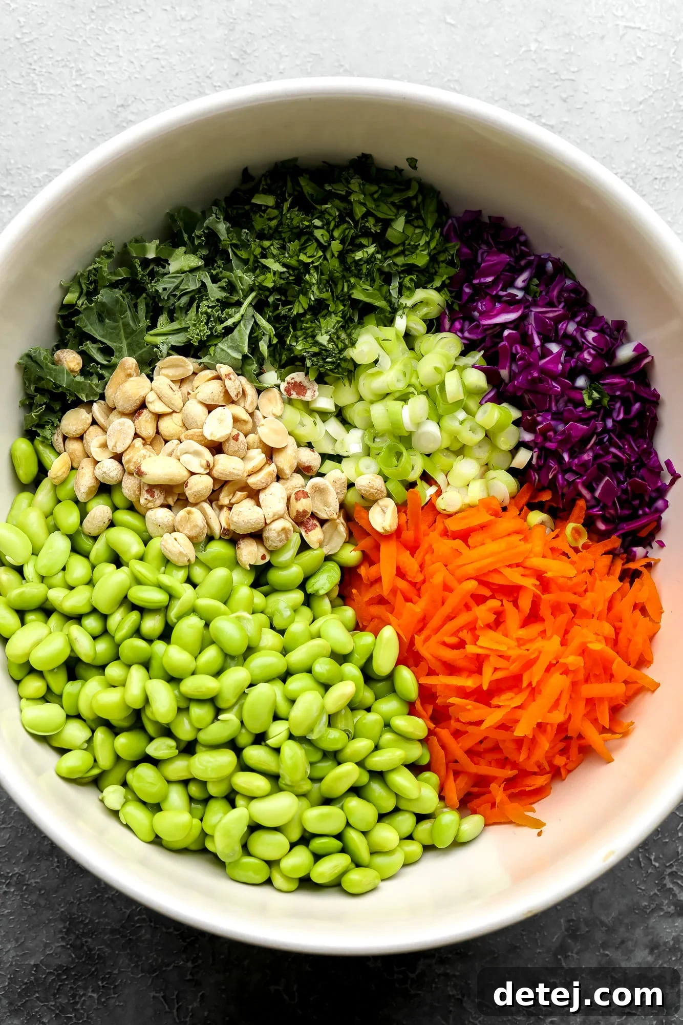 All the fresh ingredients for Edamame Salad, including vibrant edamame, shredded cabbage and carrots, kale, and green onions, gathered in a large white bowl before mixing.