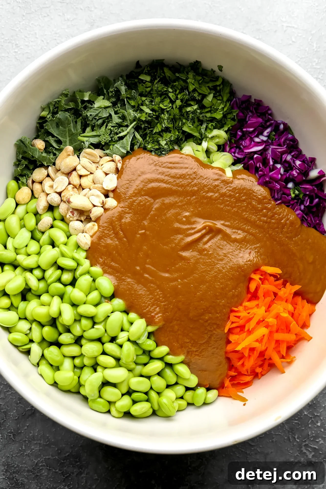 The colorful ingredients of the Edamame Salad, including edamame, vegetables, and nuts, generously coated in creamy peanut sauce within a large white bowl, ready to be chilled and served.