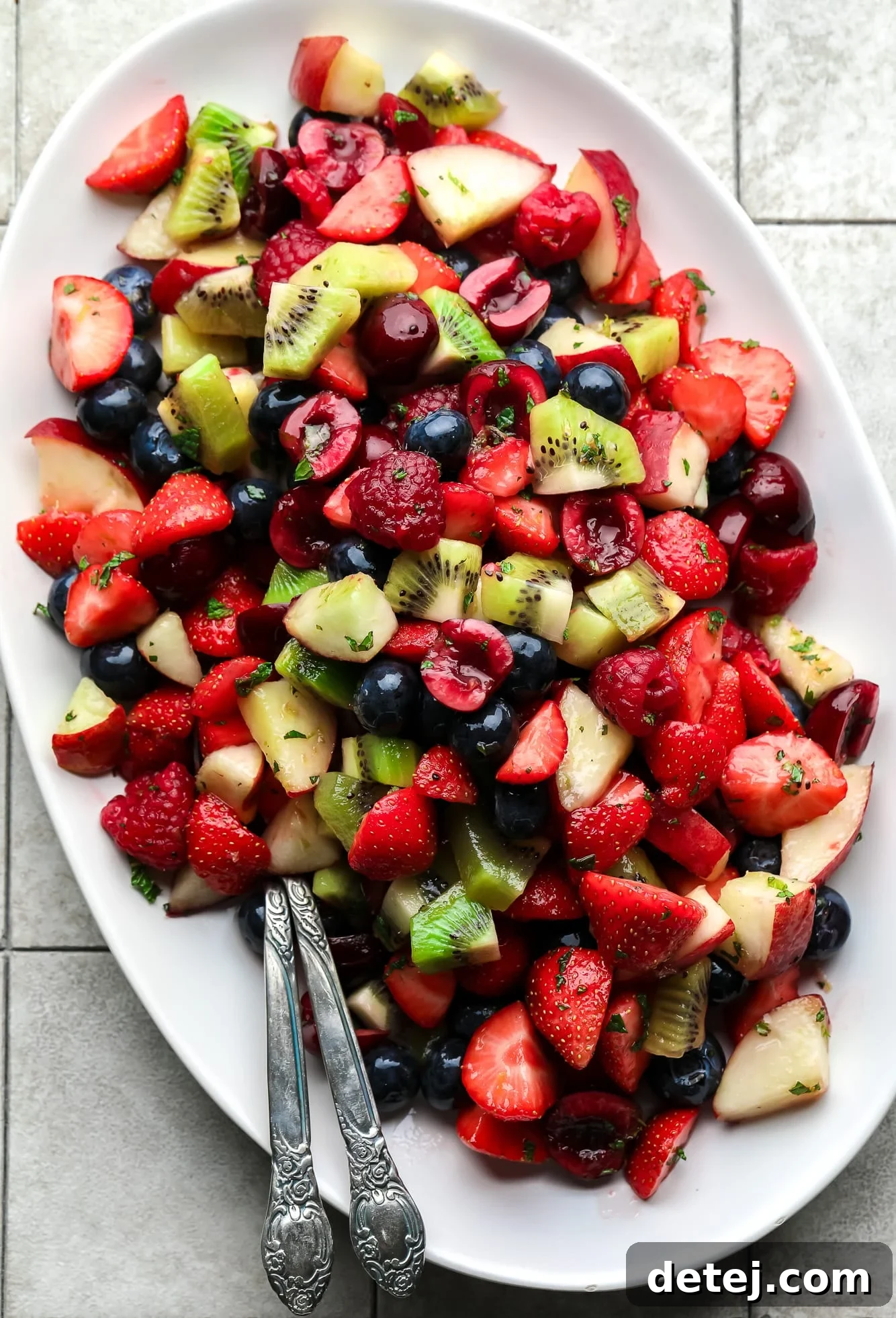 A beautifully arranged fruit salad on a pristine white serving plate, accompanied by two elegant spoons, ready to be enjoyed. The vibrant colors of fresh berries, sliced kiwi, and peaches glisten, showcasing the irresistible appeal of this summer dish.