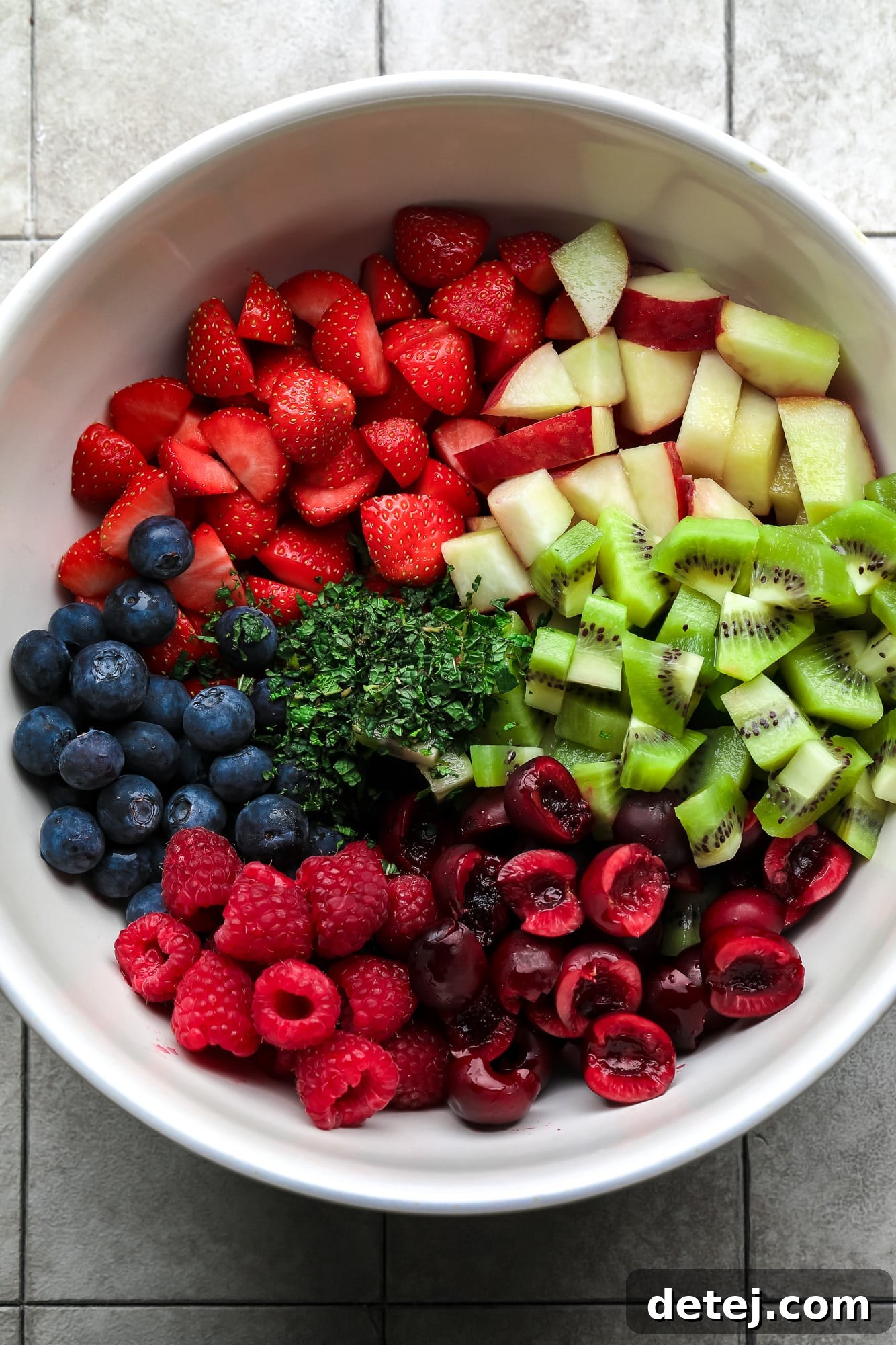 All the fresh ingredients for a fruit salad neatly arranged in a large white bowl before being mixed. You can see whole strawberries, blueberries, raspberries, cherries, sliced peaches, and kiwi, highlighting the freshness and variety of the recipe.