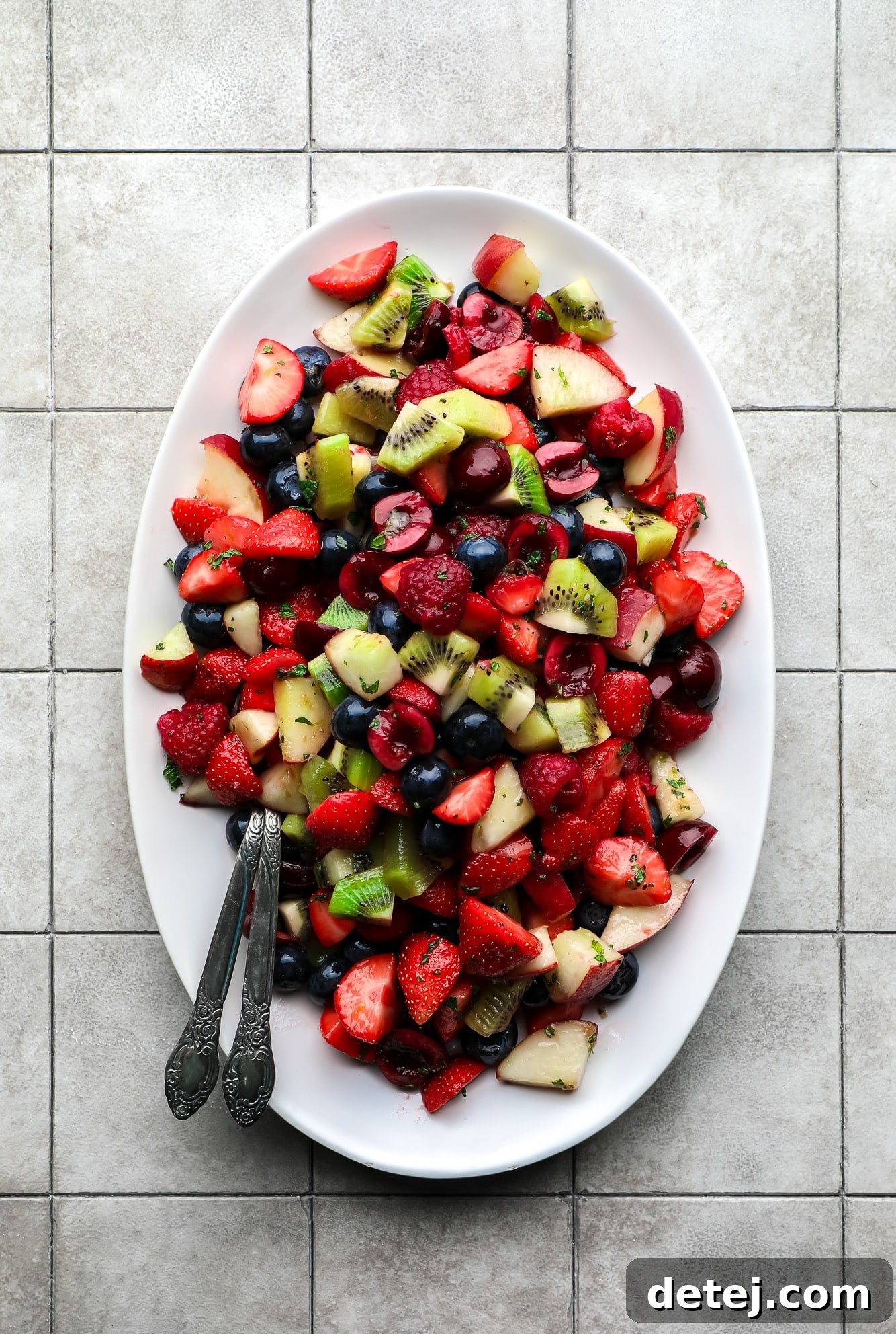 Another view of the delicious fruit salad presented on a white serving plate with two spoons, highlighting its vibrant colors and freshness, ready for enjoyment.