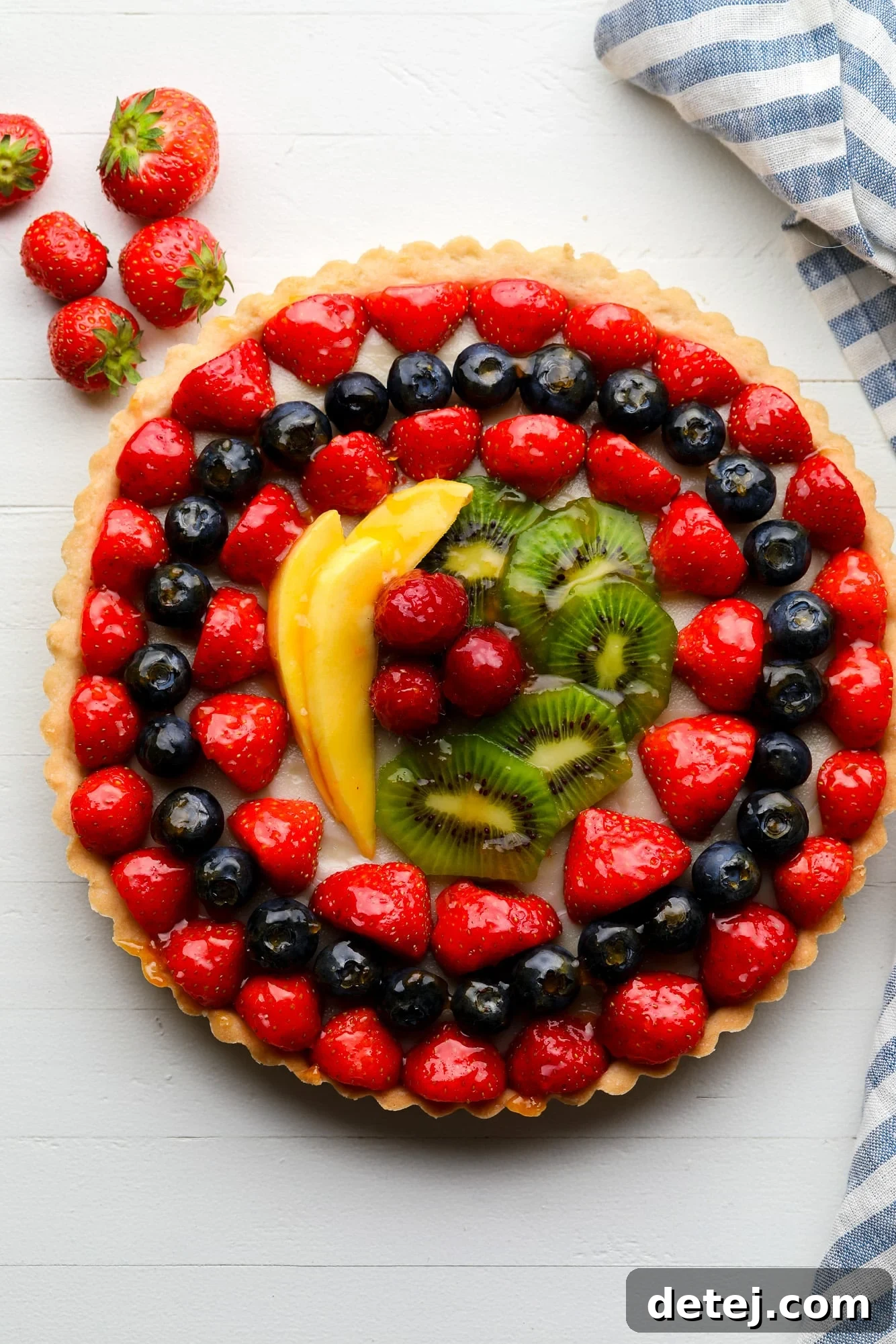 A beautifully arranged vegan fruit tart featuring rows of glazed berries, kiwi, and mango, glistening under a light glaze.