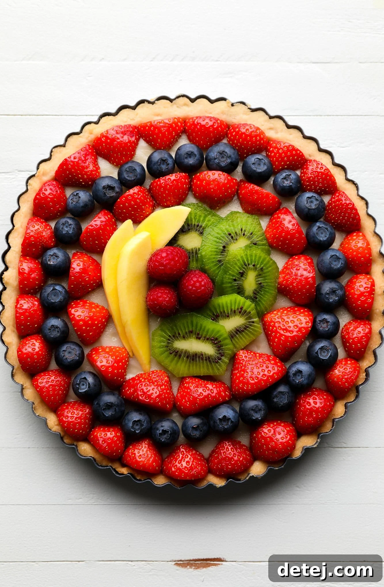 A vibrant vegan fruit tart topped with meticulously arranged rows of fresh berries, kiwi, and mango, ready to be served.