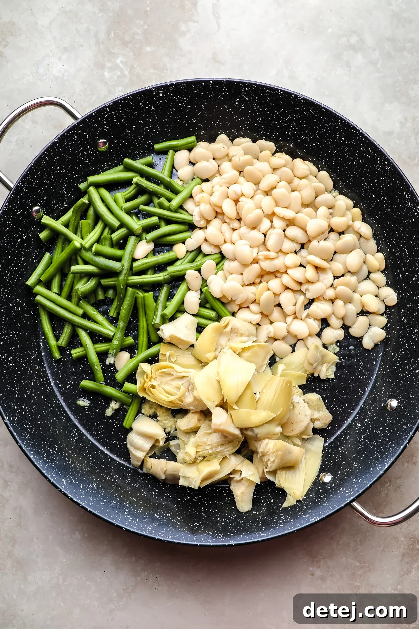 Green beans, artichoke hearts, and white beans gently sautéing in a large black paella pan.
