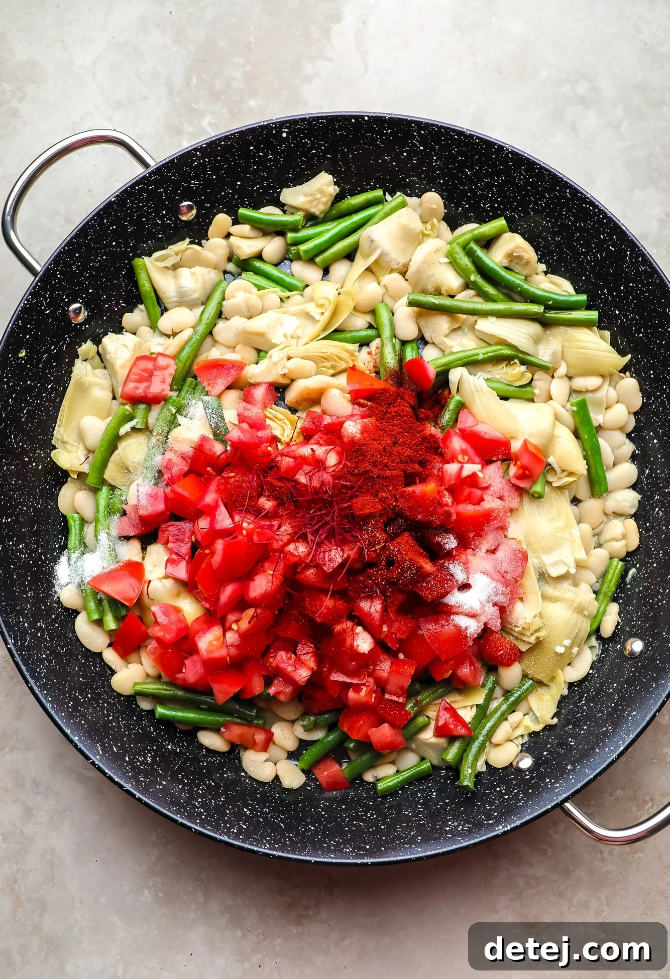 Chopped tomatoes and delicate saffron threads resting on top of beans and artichoke hearts in a black paella pan, ready for the broth.