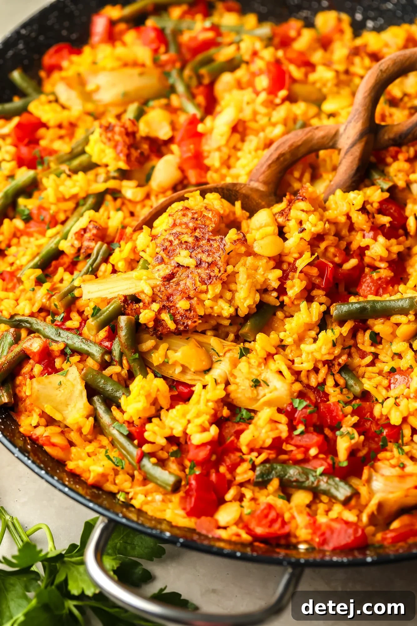 A wooden spoon delicately lifting a scoop of delicious vegan paella, showcasing the tender rice and vegetables, from a large black paella pan.
