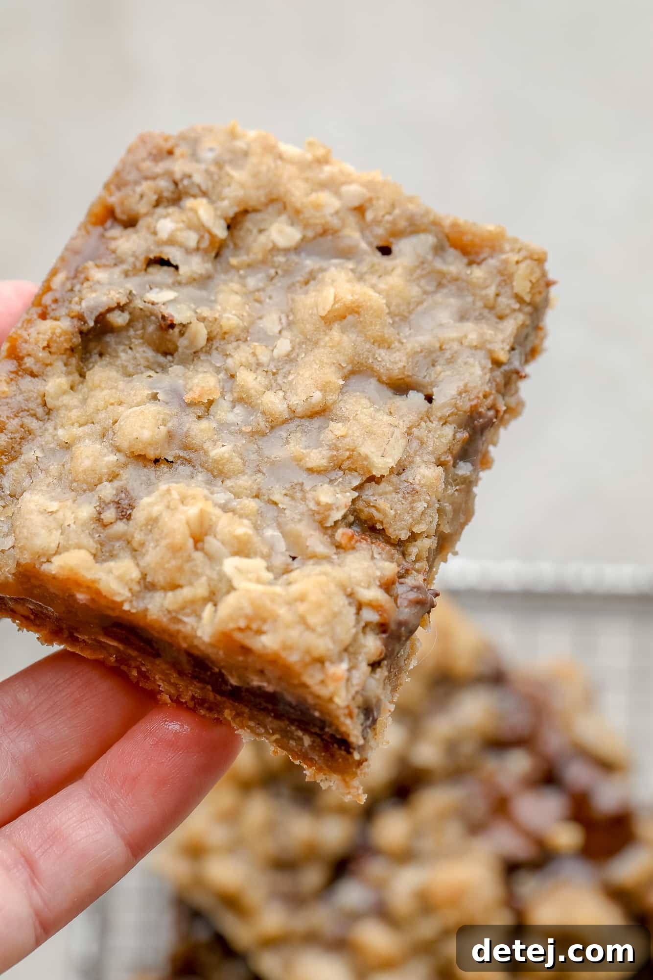A woman's hand delicately holding a single Vegan Carmelita bar, with the distinct layers of golden oatmeal crust, melted chocolate, and luscious caramel clearly visible and tempting.